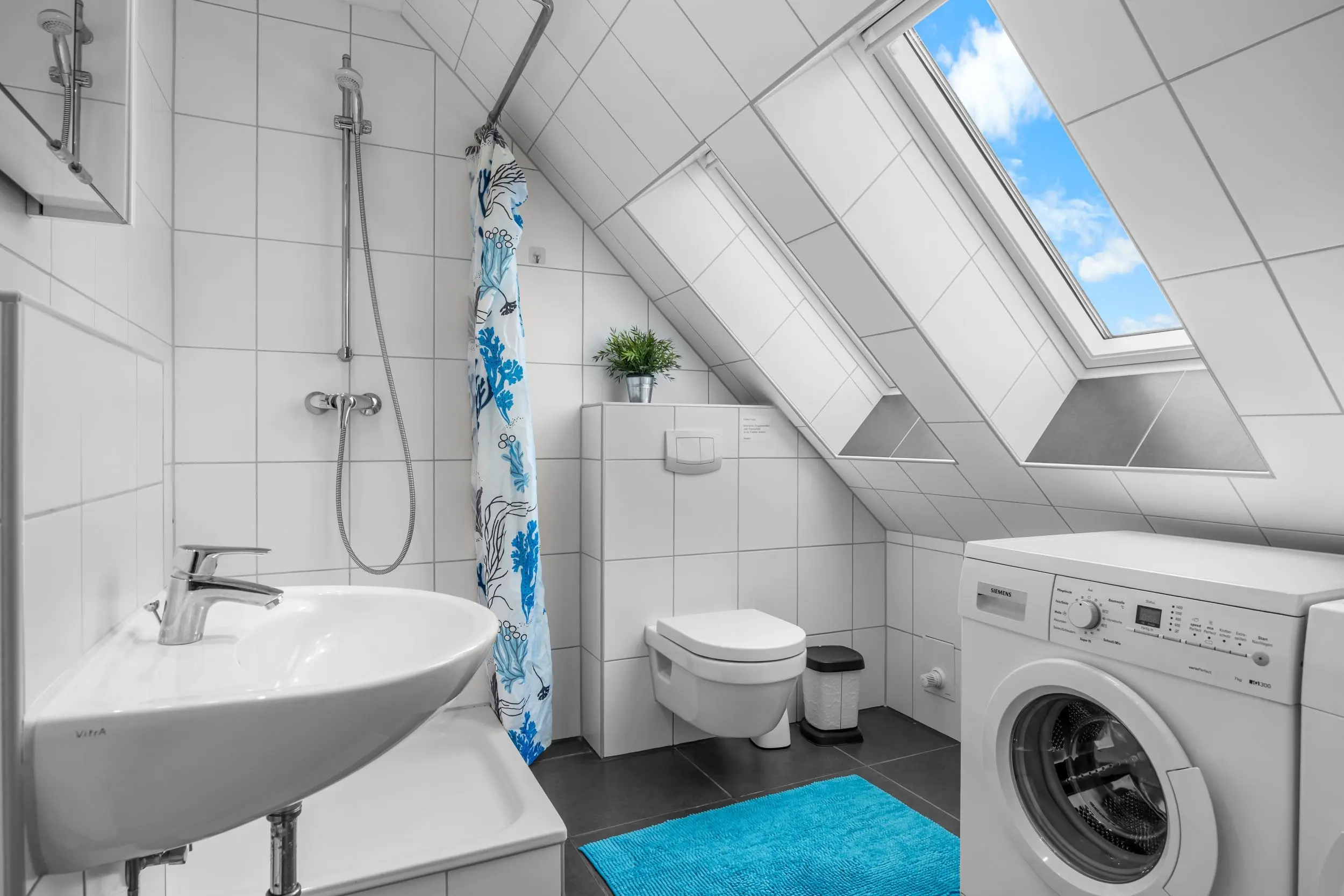 Attic bathroom with white tiles, featuring a front-loading washing machine, toilet, sink, and a shower with a blue-pattern curtain under a slanted ceiling and a skylight showing blue sky.