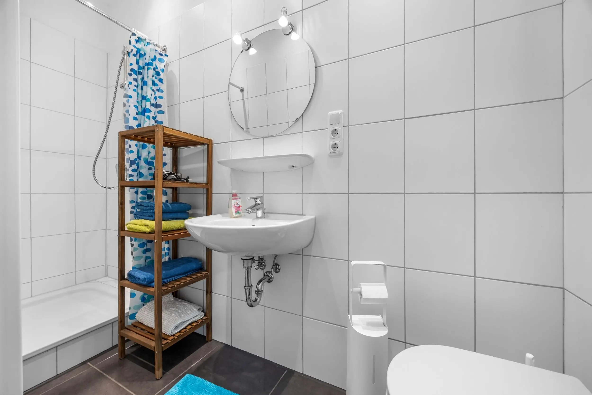 Bathroom with white tiled walls, round mirror above a white sink, blue patterned shower curtain, and a wooden shelf holding towels nearby