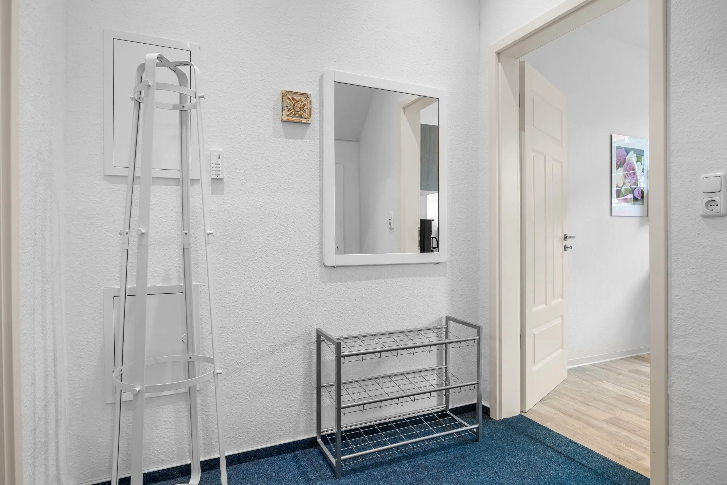 White-walled hallway with a tall folding ladder, a metal rack, and a wall mirror near a door; a framed flower print hangs on the wall.