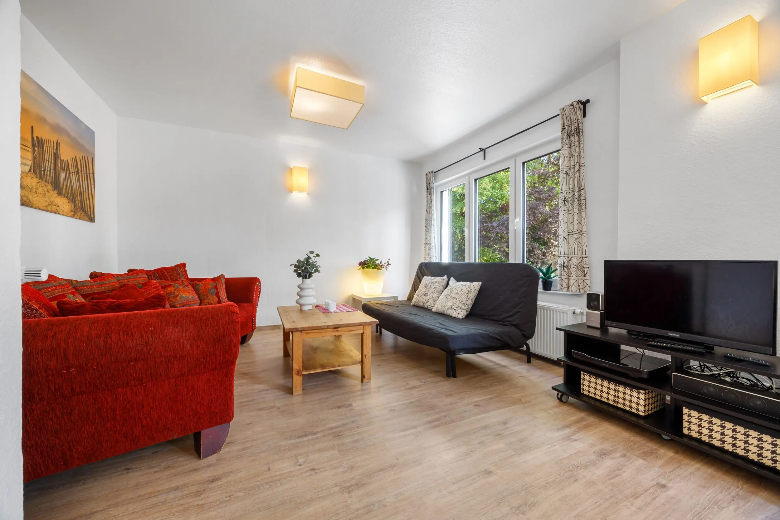 Cozy living room with a red sofa, black futon, and wooden coffee table by a large window with patterned curtains.