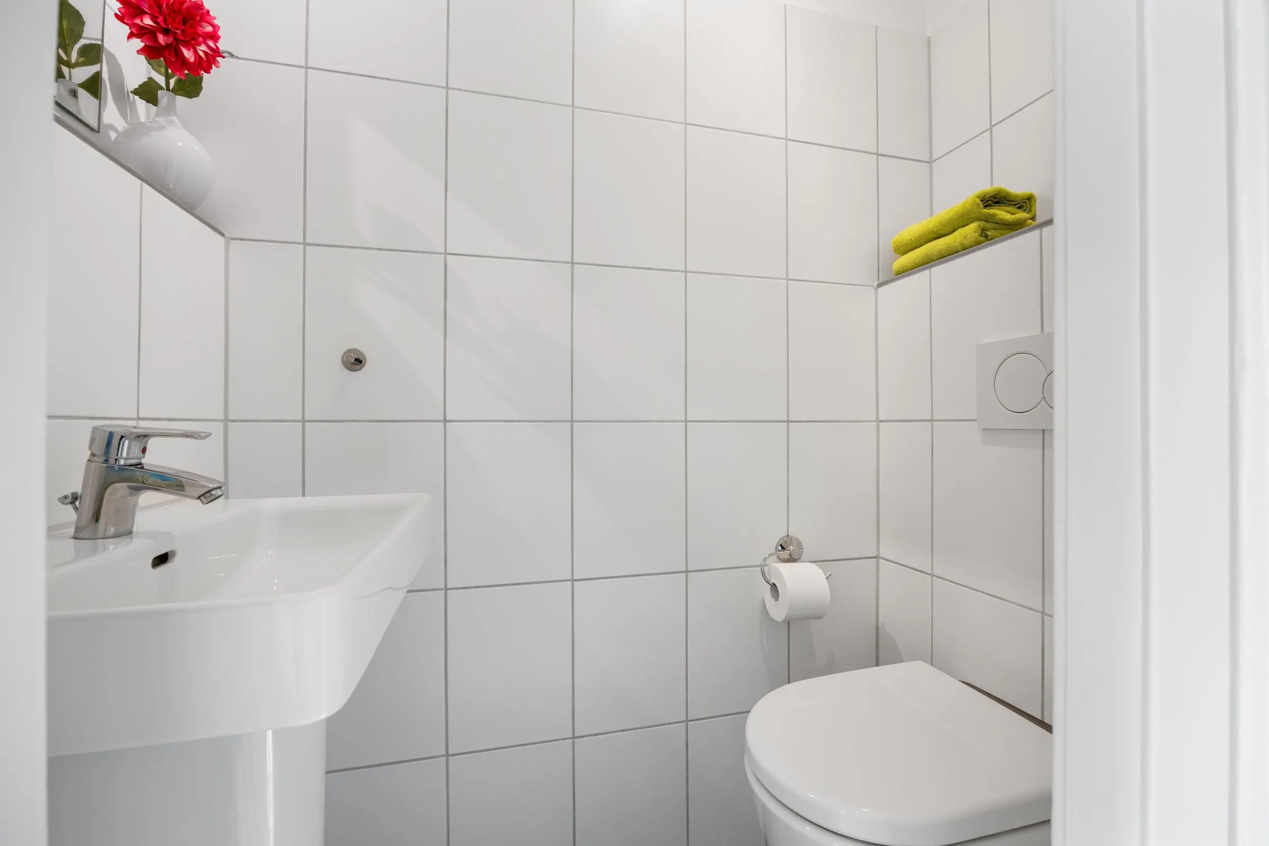 White tiled bathroom with a white pedestal sink, chrome faucet, and a wall shelf holding a vase of bright red flowers; toilet and toilet paper holder nearby.