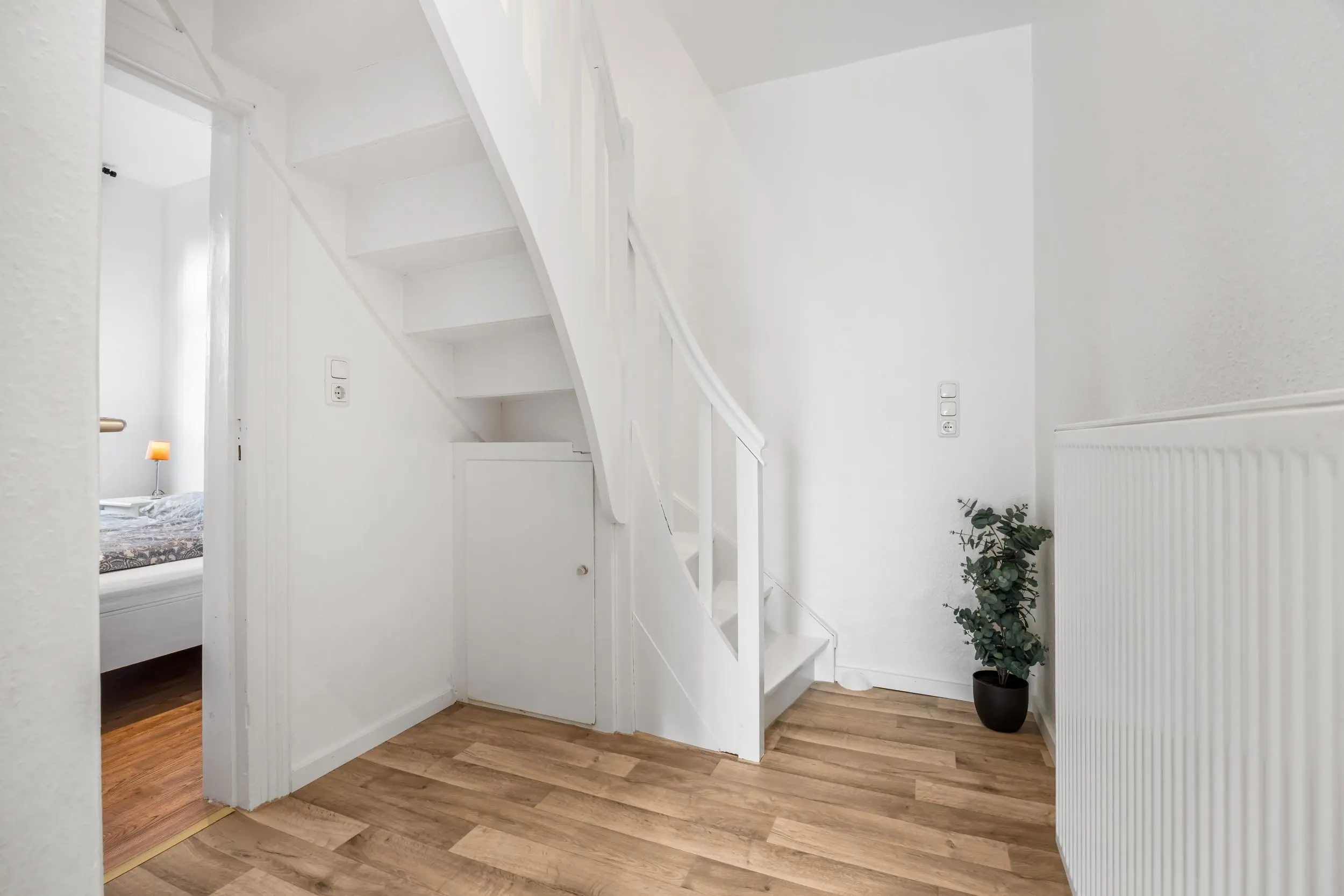 Bright white interior hallway with a narrow white staircase, radiator on the right, and a potted plant; doorway reveals a bedroom with a lamp and bed.