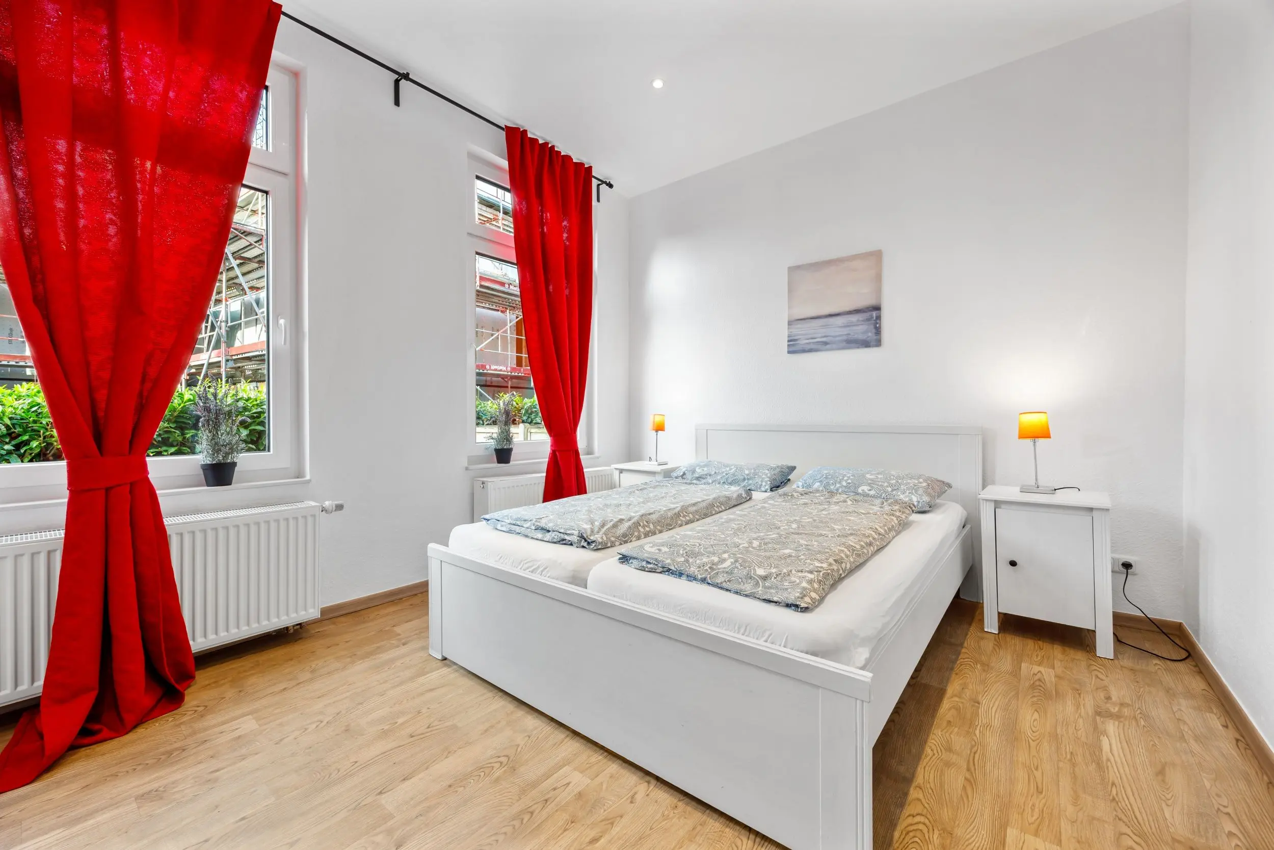 Bright bedroom with two single beds, white furniture, red floor-length curtains, and a seascape painting above the headboard.