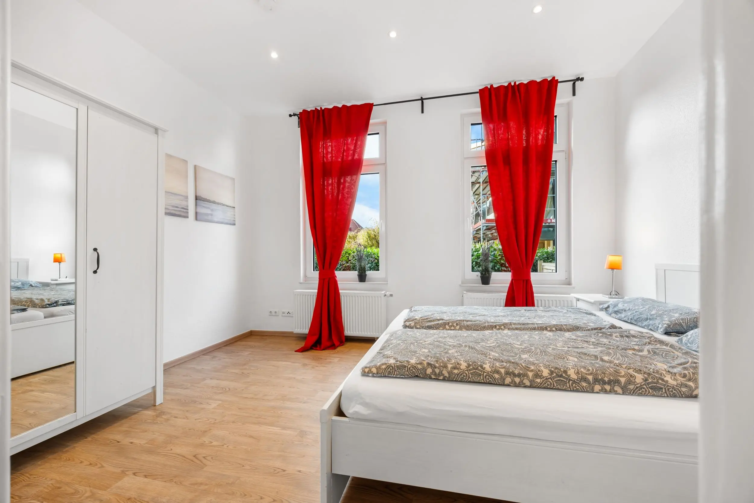 Bright white bedroom with red curtains framing two windows and two beds with patterned gray bedding in the foreground.