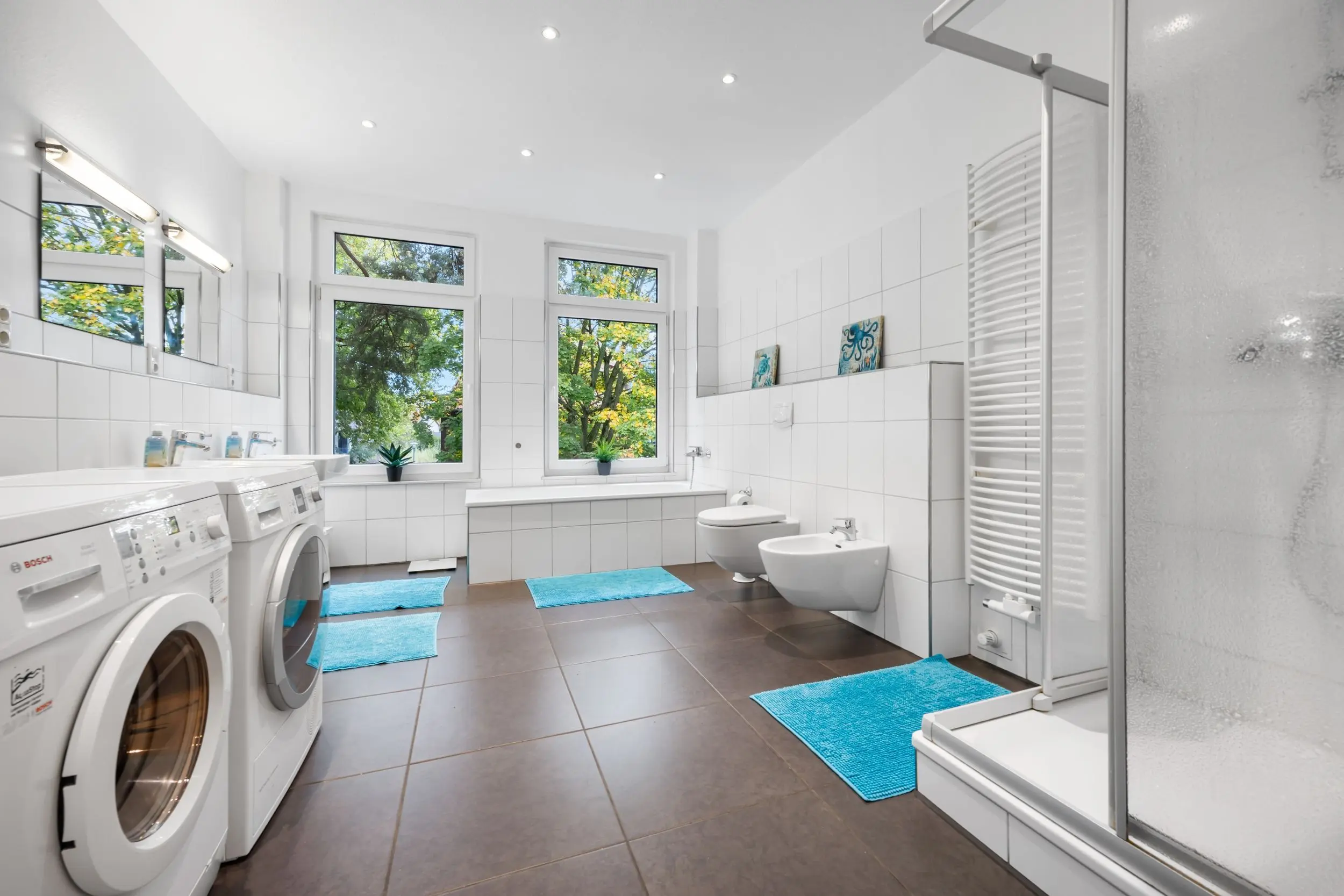 Bright white bathroom with two front-loading washing machines, large windows, and turquoise bath mats.