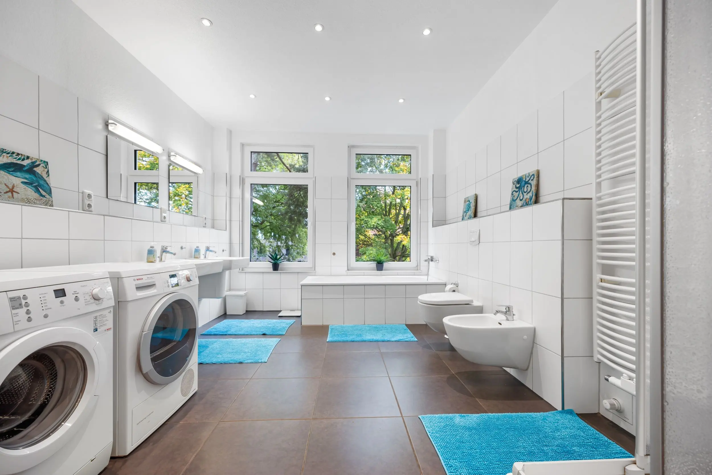 Bright white bathroom with washing machines along left, two sinks, bidet and toilet, large windows with greenery outside, and blue bath mats on a tiled floor.