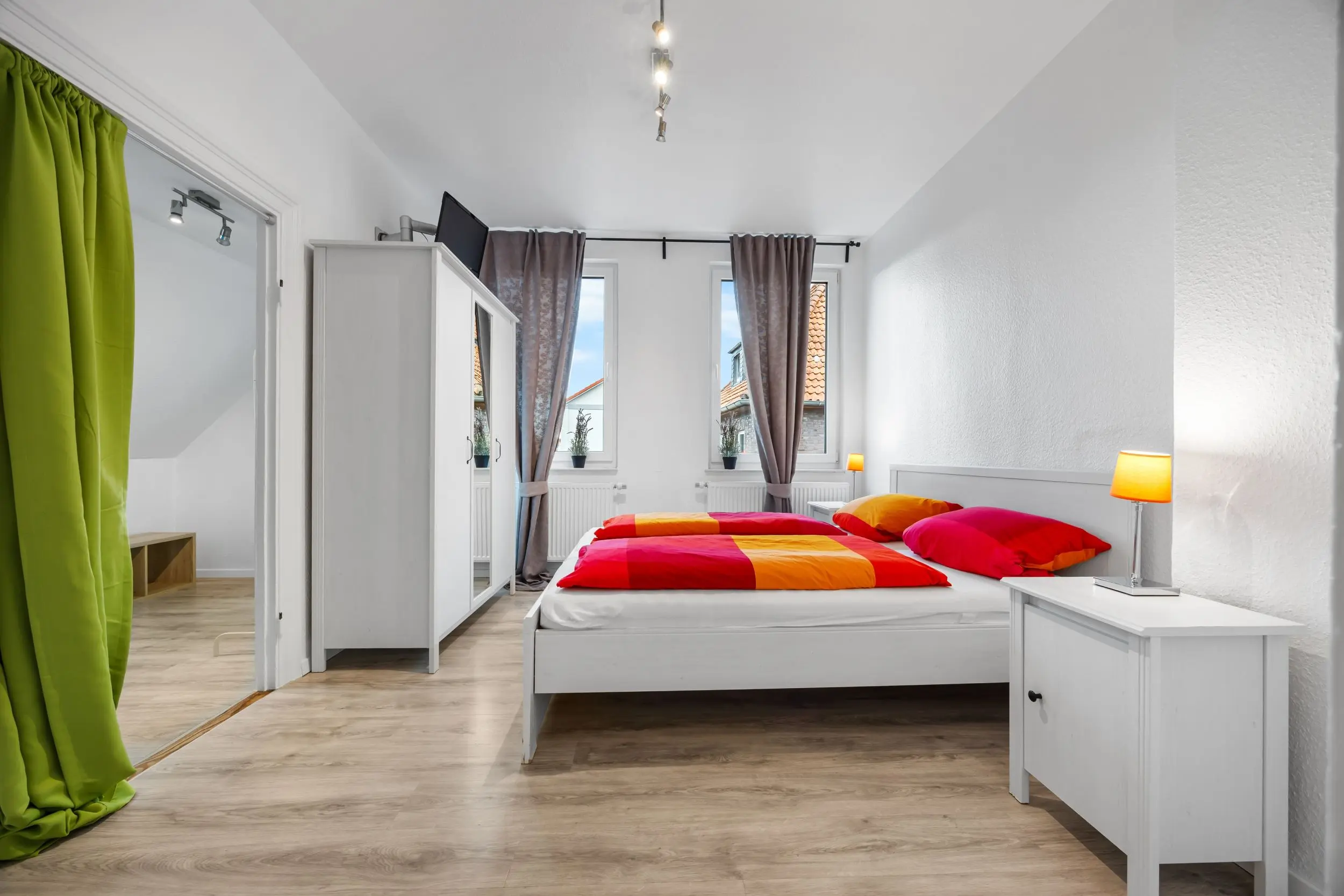 Bright, modern bedroom with two single beds in orange-pink duvets, white furniture and gray curtains by two windows.