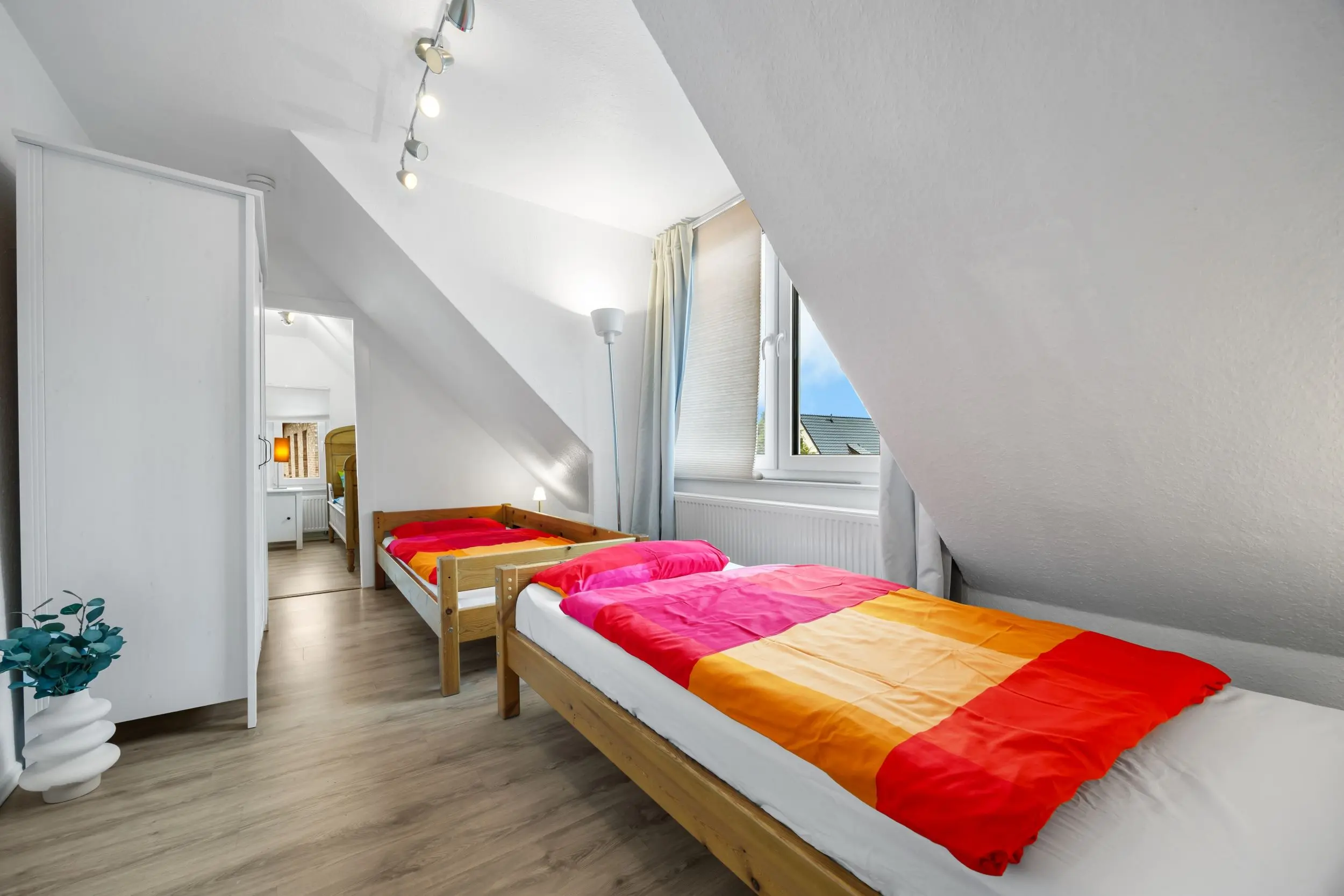Bright attic bedroom with two wooden single beds finished in orange-pink bedding under a slanted ceiling, window on the right, white wardrobe left, and a potted plant nearby