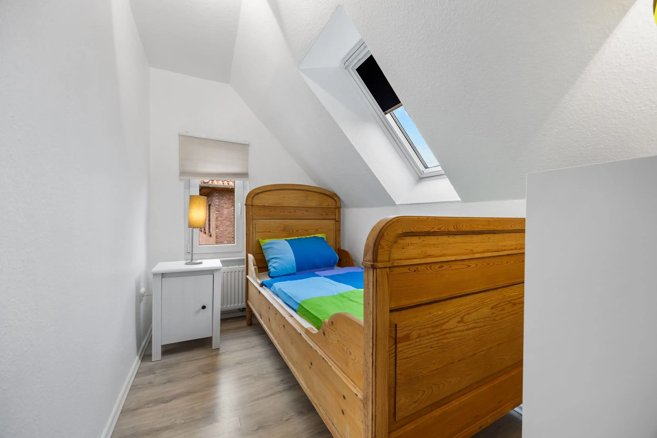 Attic bedroom with sloped white walls, two wooden single beds, blue-green bedding, a white bedside table and lamp, and a skylight.