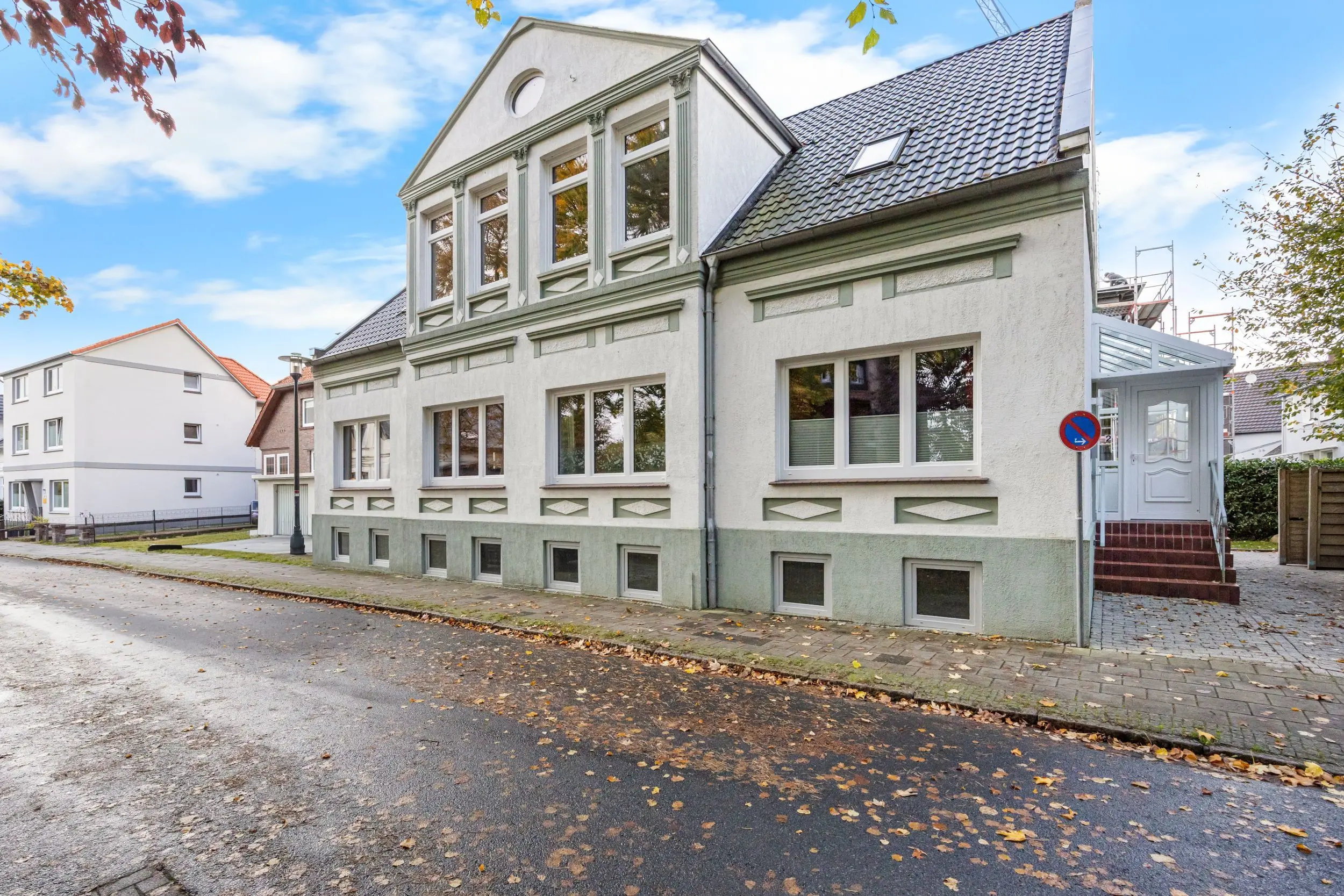 Two-story gray building with decorative window frames along a sunny street; autumn leaves scattered on the pavement.