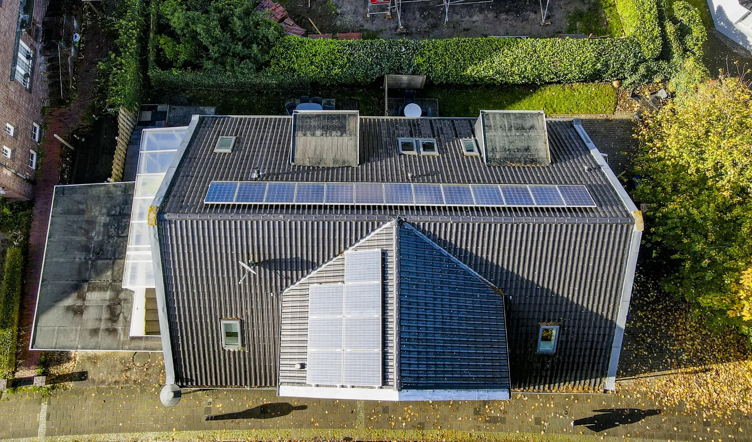 Top-down aerial view of a house roof with a row of solar panels, surrounded by hedges, trees, and a paved yard area.