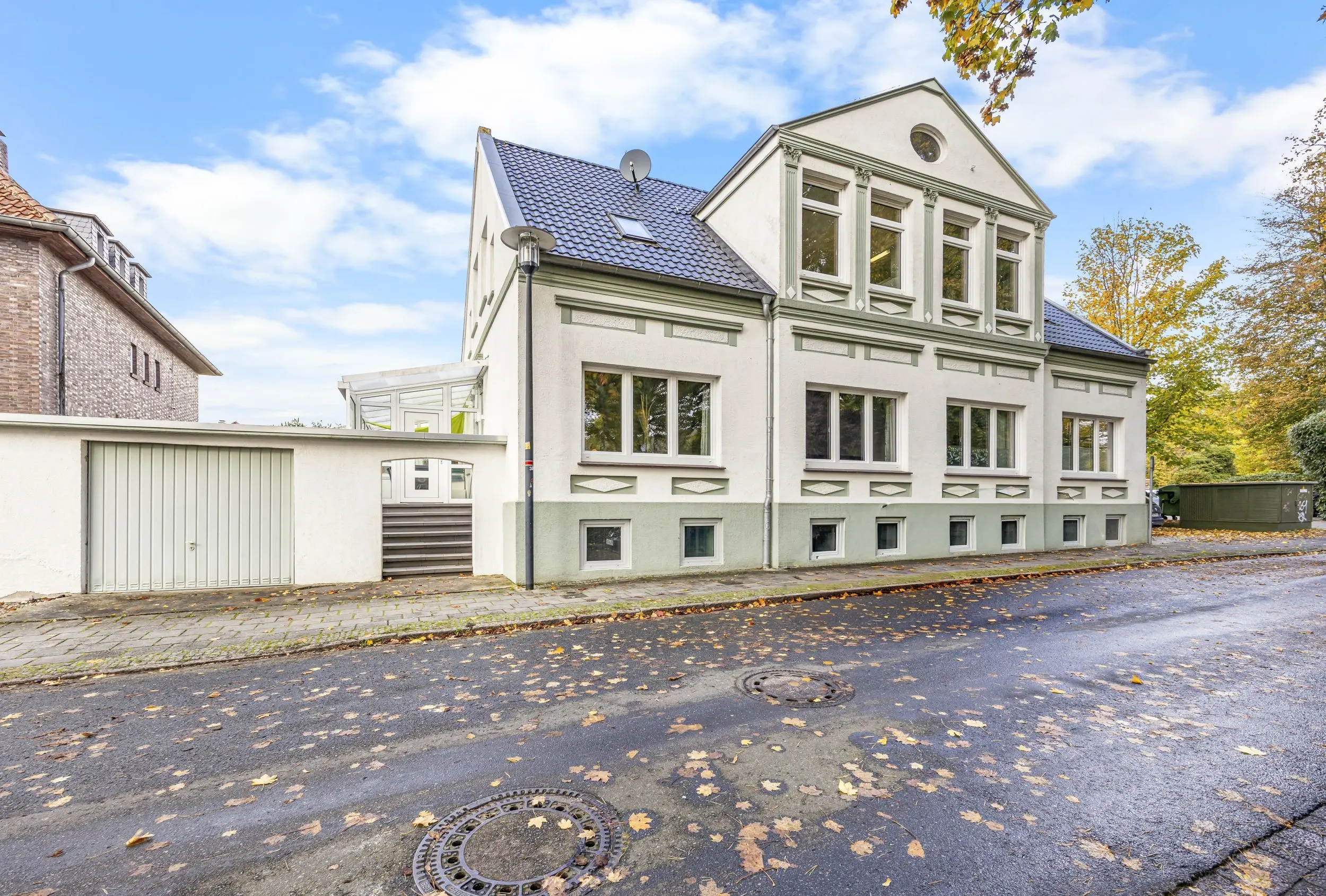 White three-story building with decorative green trim on a tree-lined street, fallen autumn leaves on the pavement.
