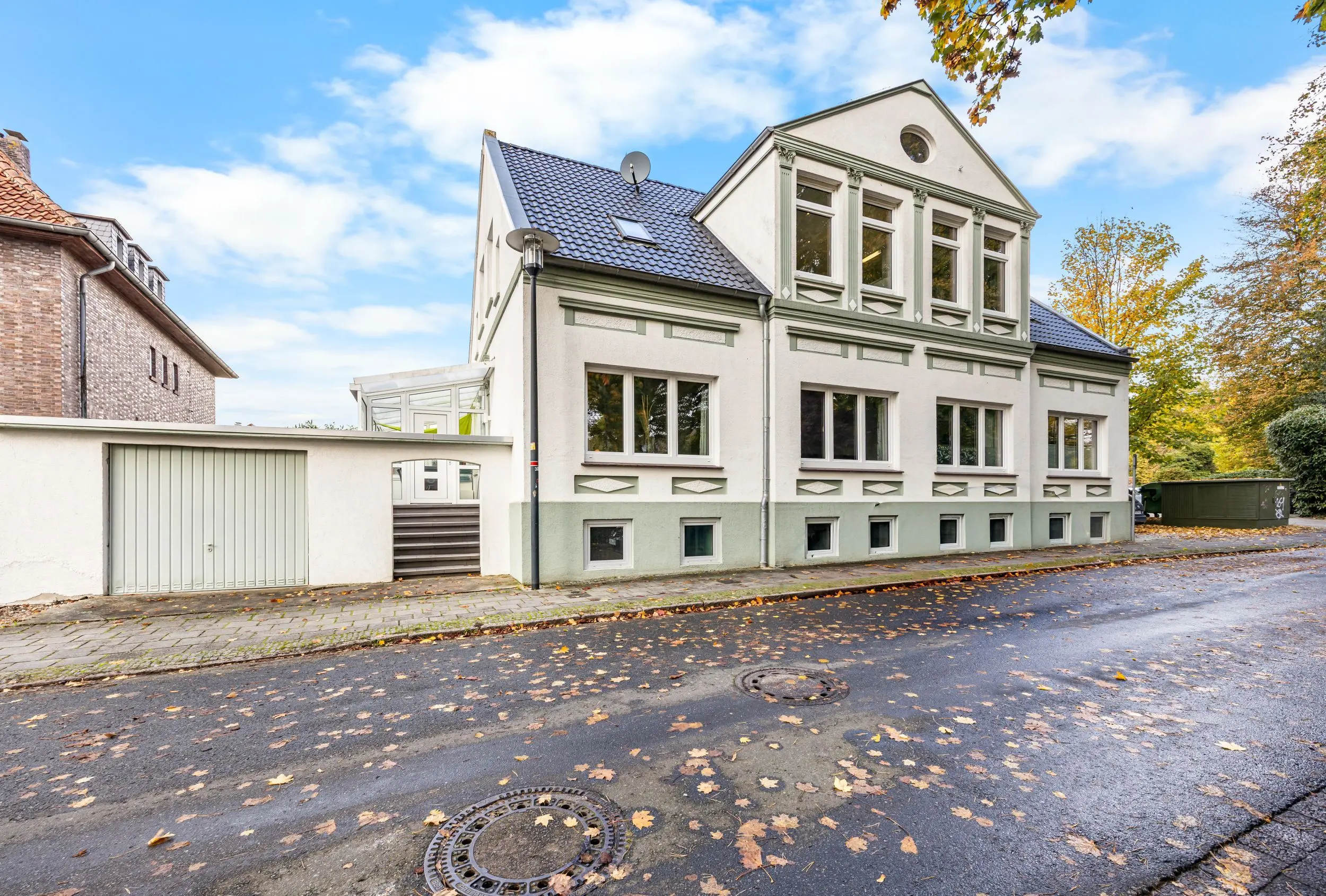 White historic townhouse with ornate green trim and a blue-tiled roof on a tree-lined autumn street; leaves scattered on the pavement.