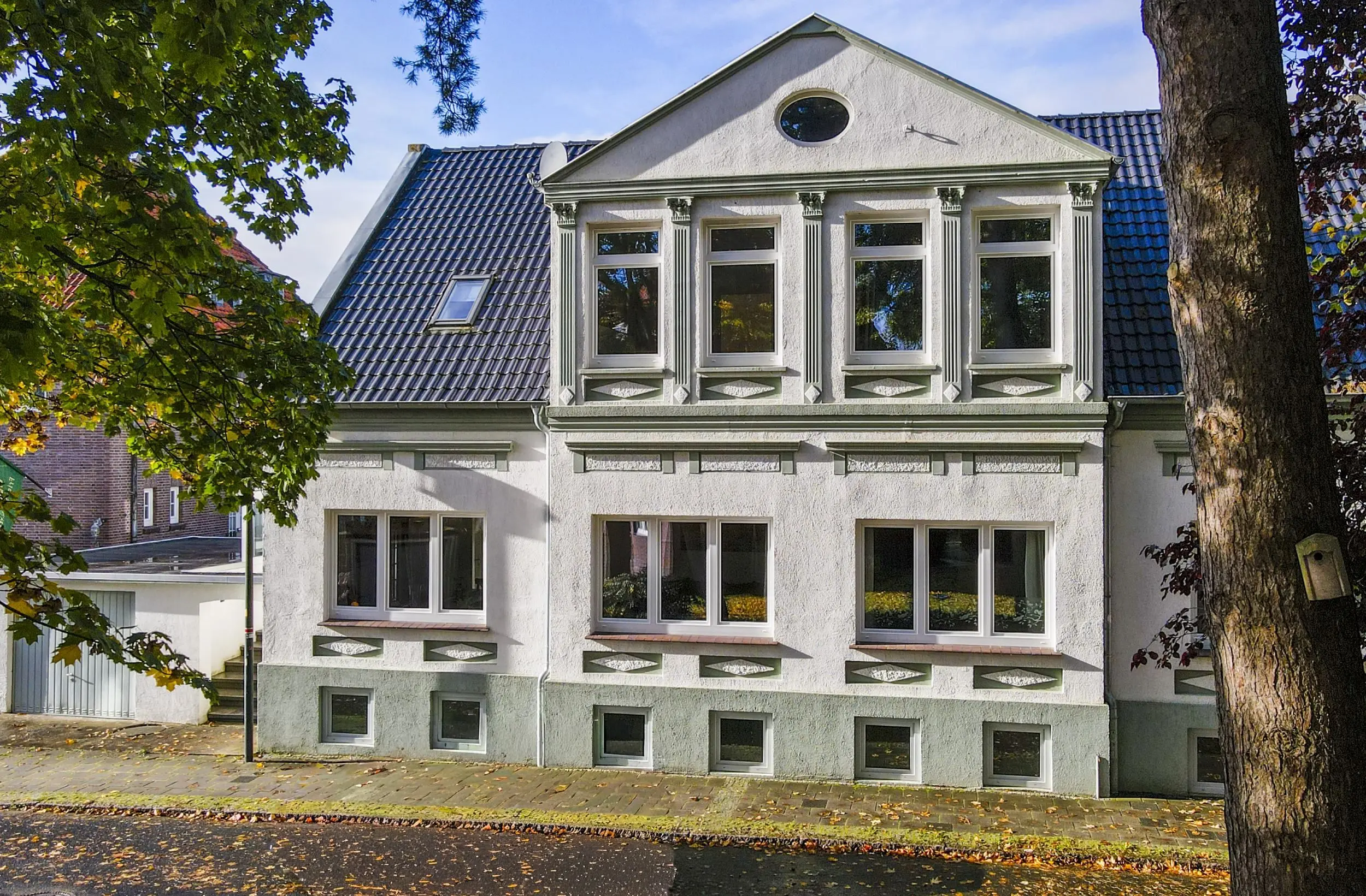White stucco townhouse with a decorative second-floor bay of four windows and green trim, brick sidewalk and trees framing the scene.