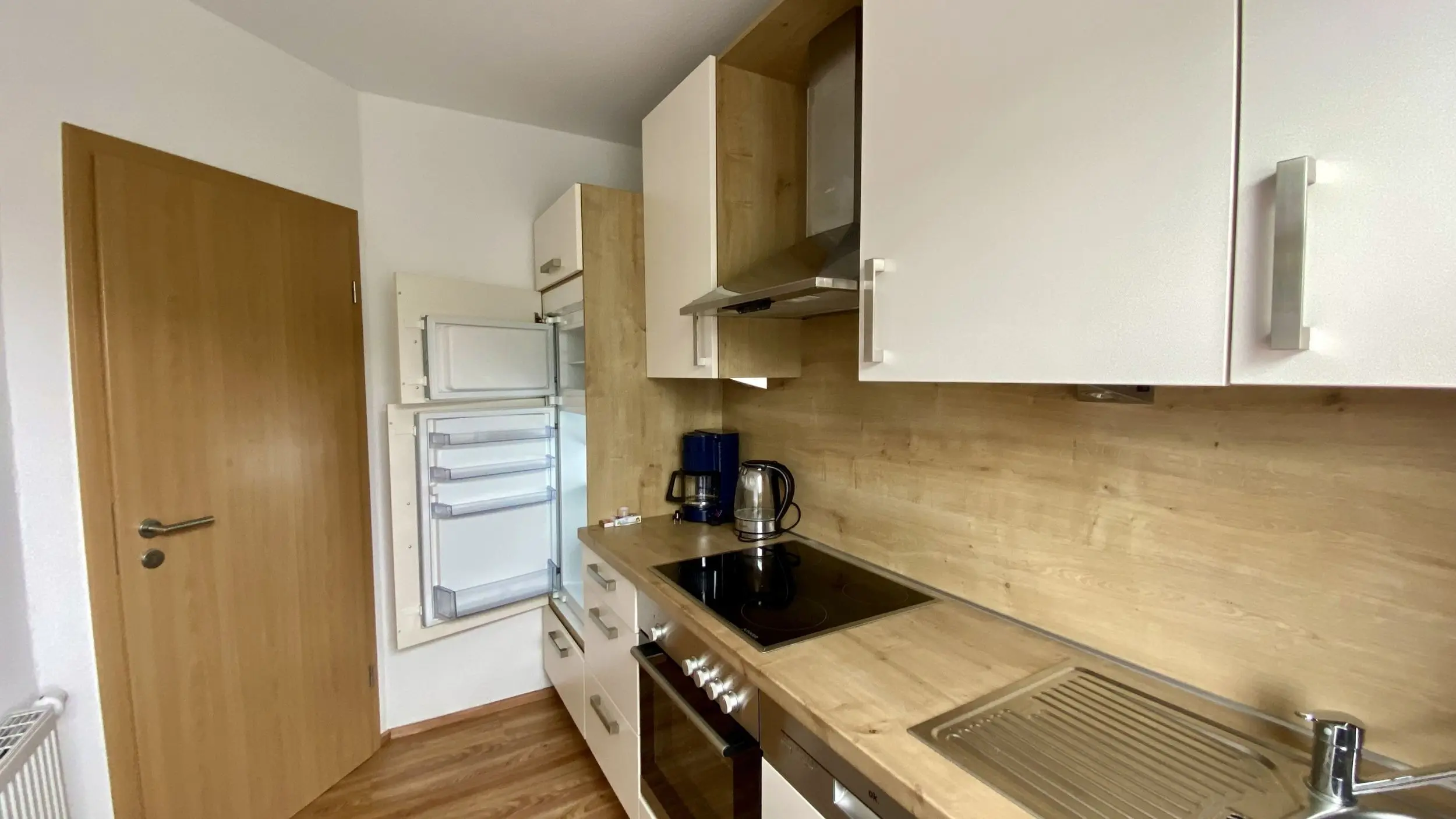 Compact modern kitchen with white upper cabinets, wood countertops, and stainless steel stove and hood; coffee maker and kettle on the counter.