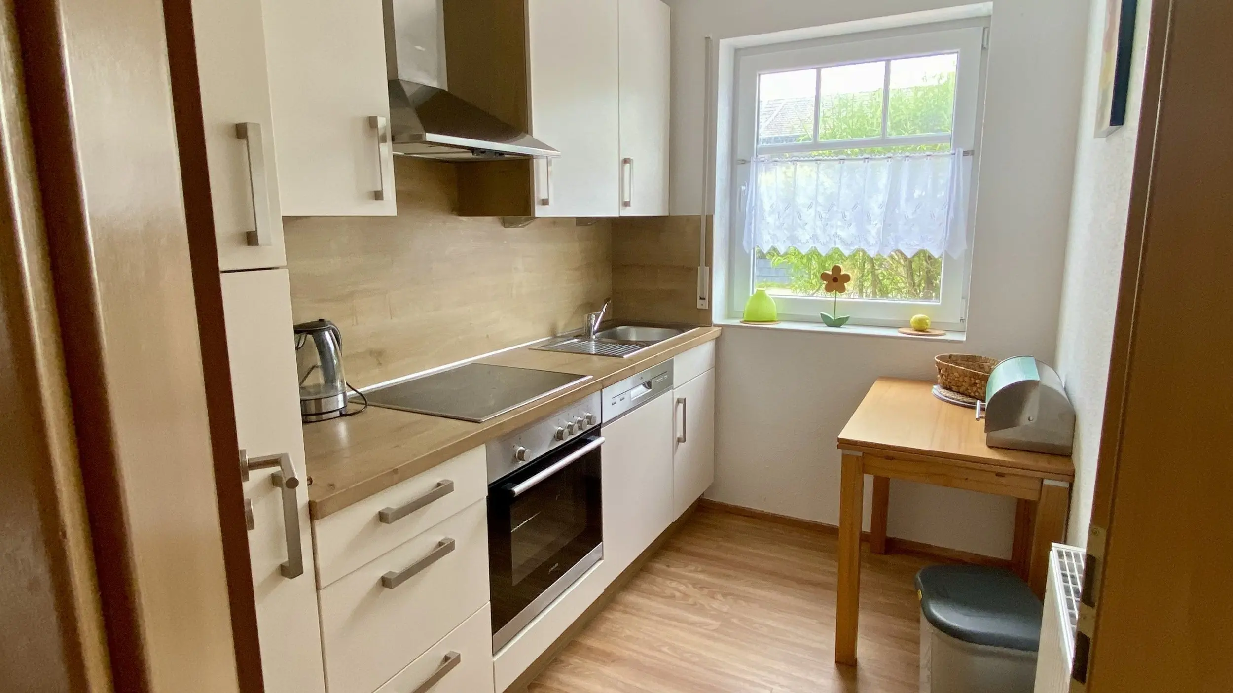 Narrow modern kitchen with white cabinets, stainless steel oven and hob, and a window with a lace curtain above the sink area.