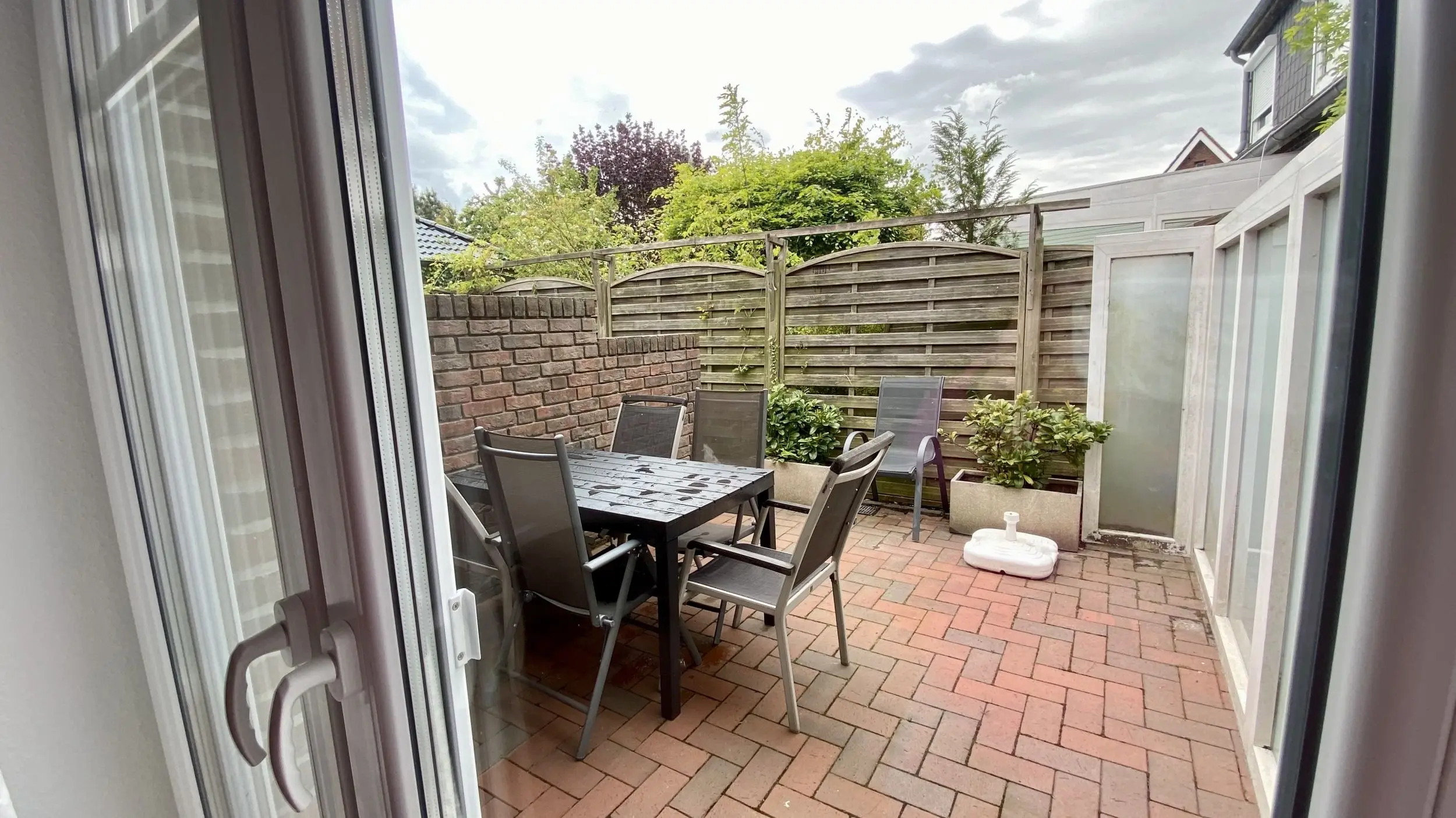 Small outdoor patio with a metal table and six chairs, brick paving, and a wooden fence, viewed from a sliding door.