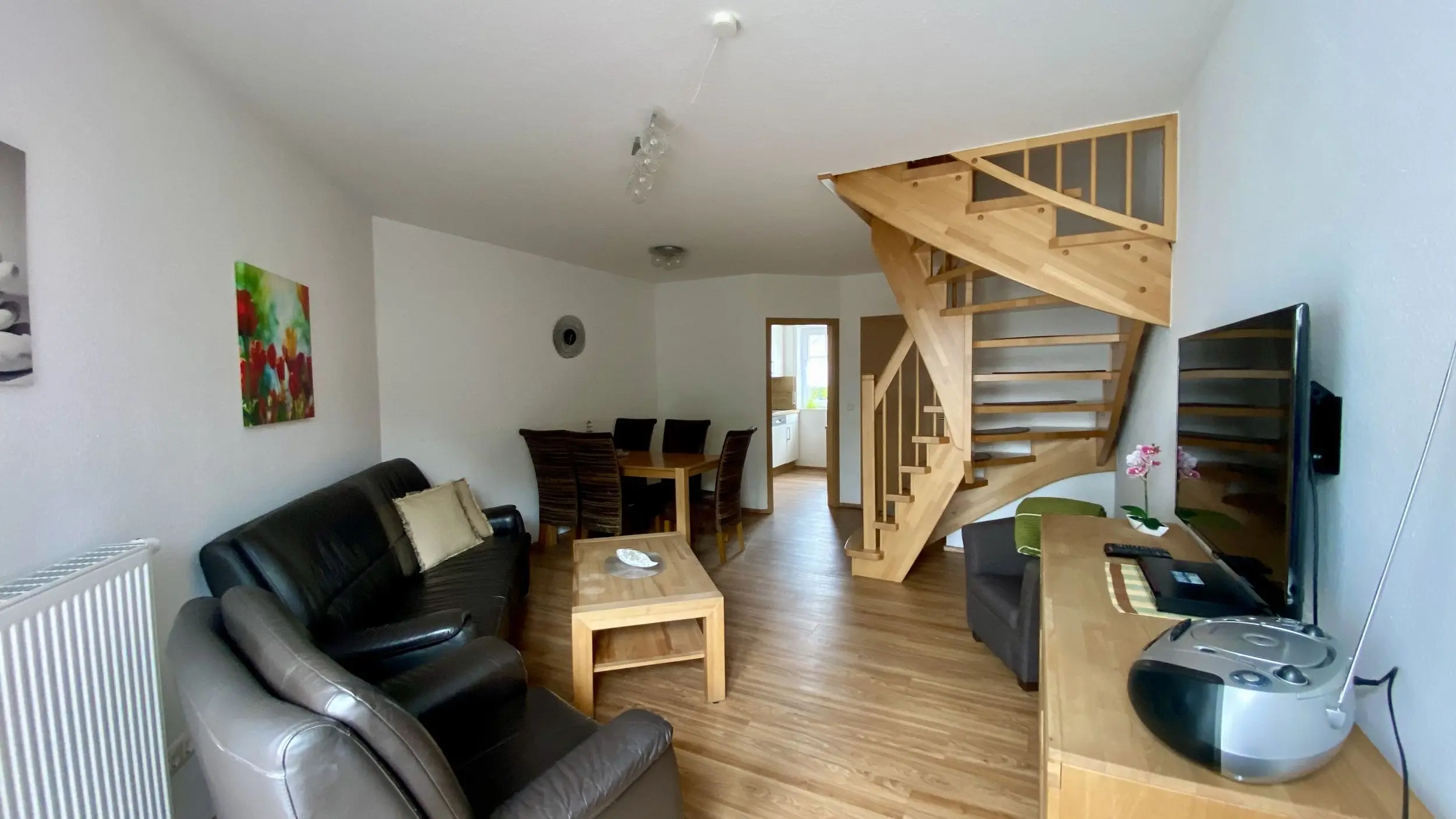 Open-plan living room with a wooden staircase, leather sofa, coffee table, dining table and chairs, and a wall-mounted TV on the right.