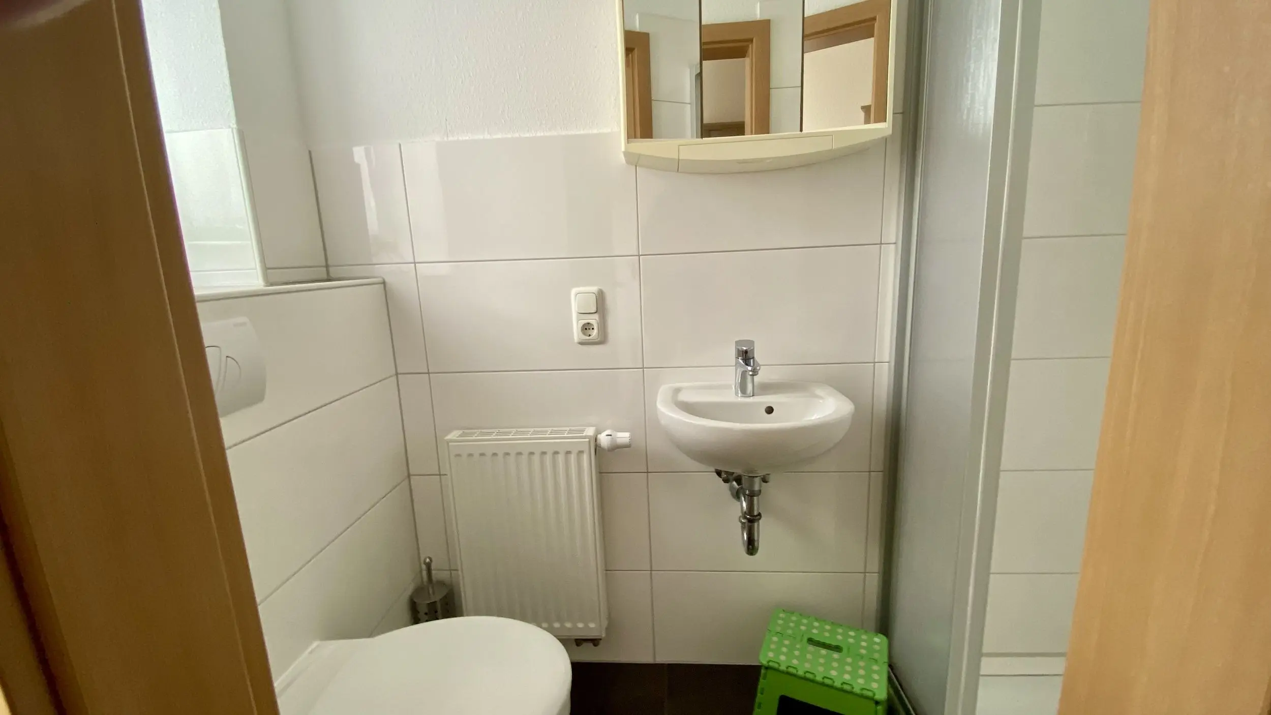 Small tiled bathroom with a white wall-mounted sink, a white toilet in the foreground, and a wall mirror cabinet above the sink; a white radiator and a green polka-dotted step stool are on the floor.
