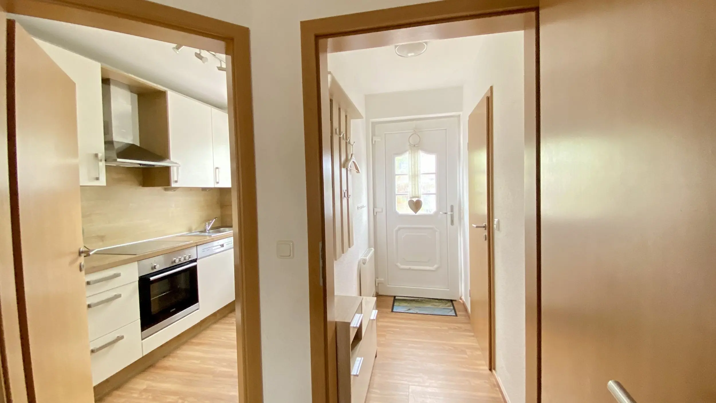 View through a doorway into a modern kitchen with white cabinets and stainless steel appliances on the left, hallway ahead with a white front door and heart ornament hanging outside the door frame.