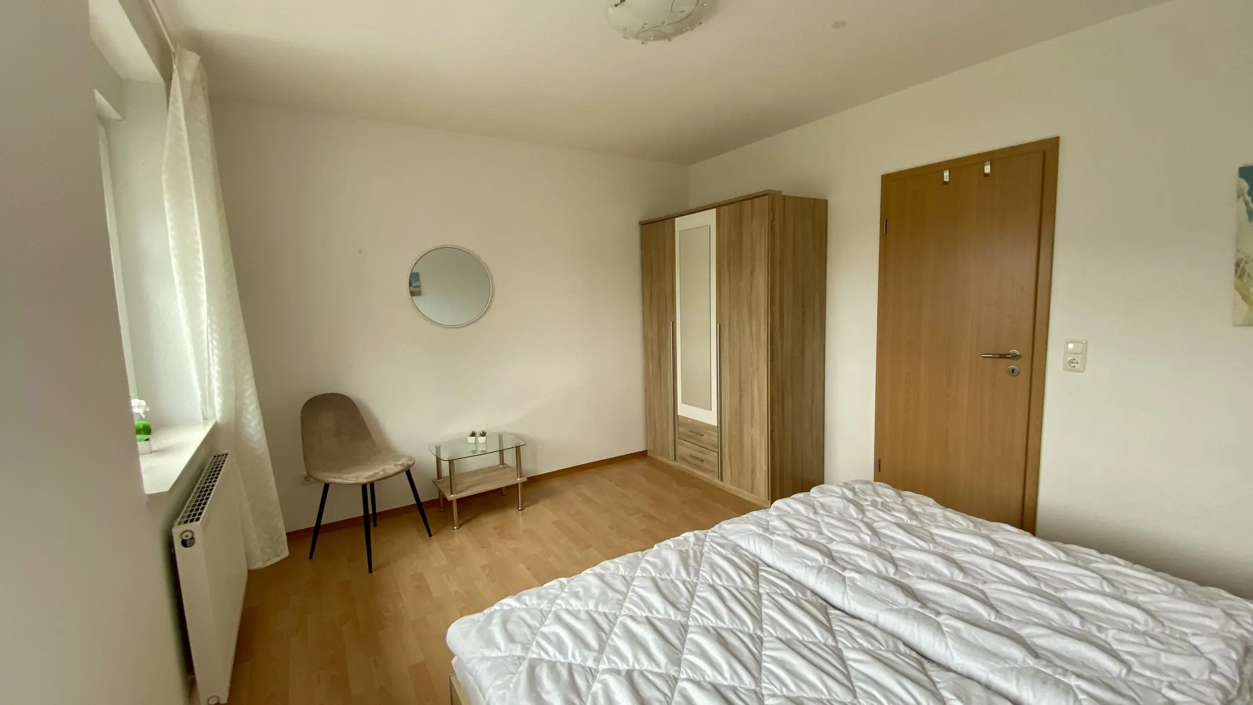 A tidy bedroom with a white quilted bed in the foreground, a wooden wardrobe, a round wall mirror, and a small glass coffee table beside a beige chair.