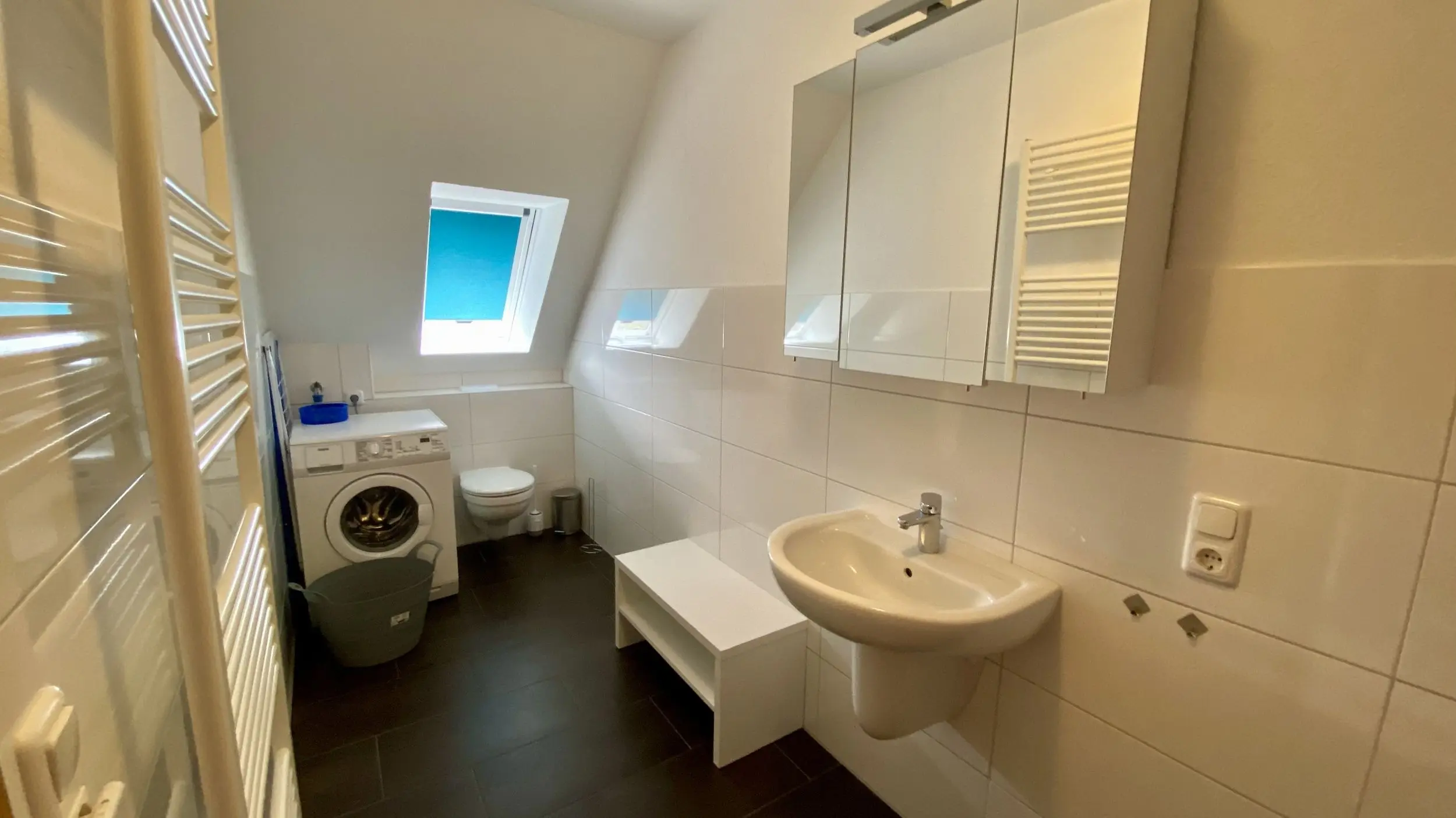 Small white-tiled bathroom with a front-loading washing machine under a skylight window, a wall mirror cabinet, a wall-mounted sink, and a white towel radiator.