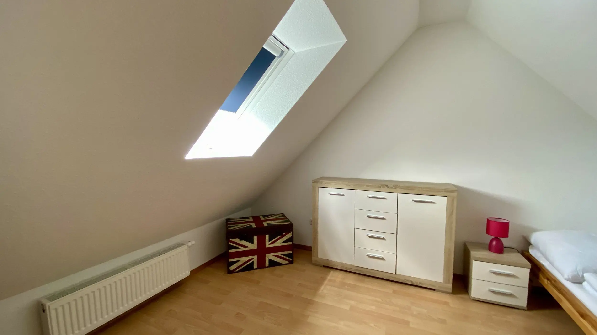 Bedroom with a slanted ceiling and a skylight window, a light wood dresser, and a small nightstand with a pink lamp. Union Jack storage boxes sit by the radiator and a bed is visible on the right.