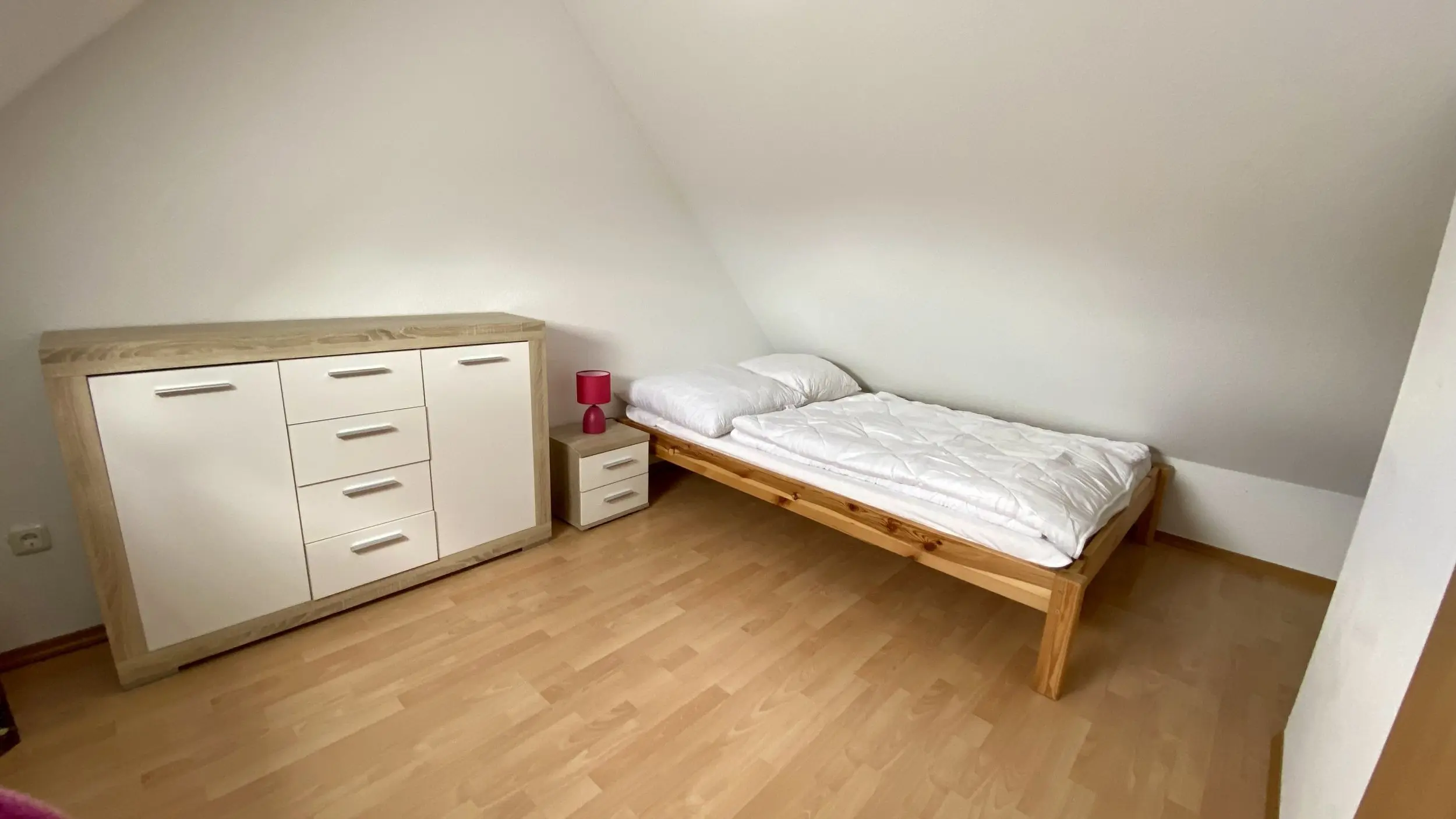 Minimalist attic bedroom with a single wooden bed, white bedding, a small nightstand with a pink lamp, and a wood dresser on a light floor.