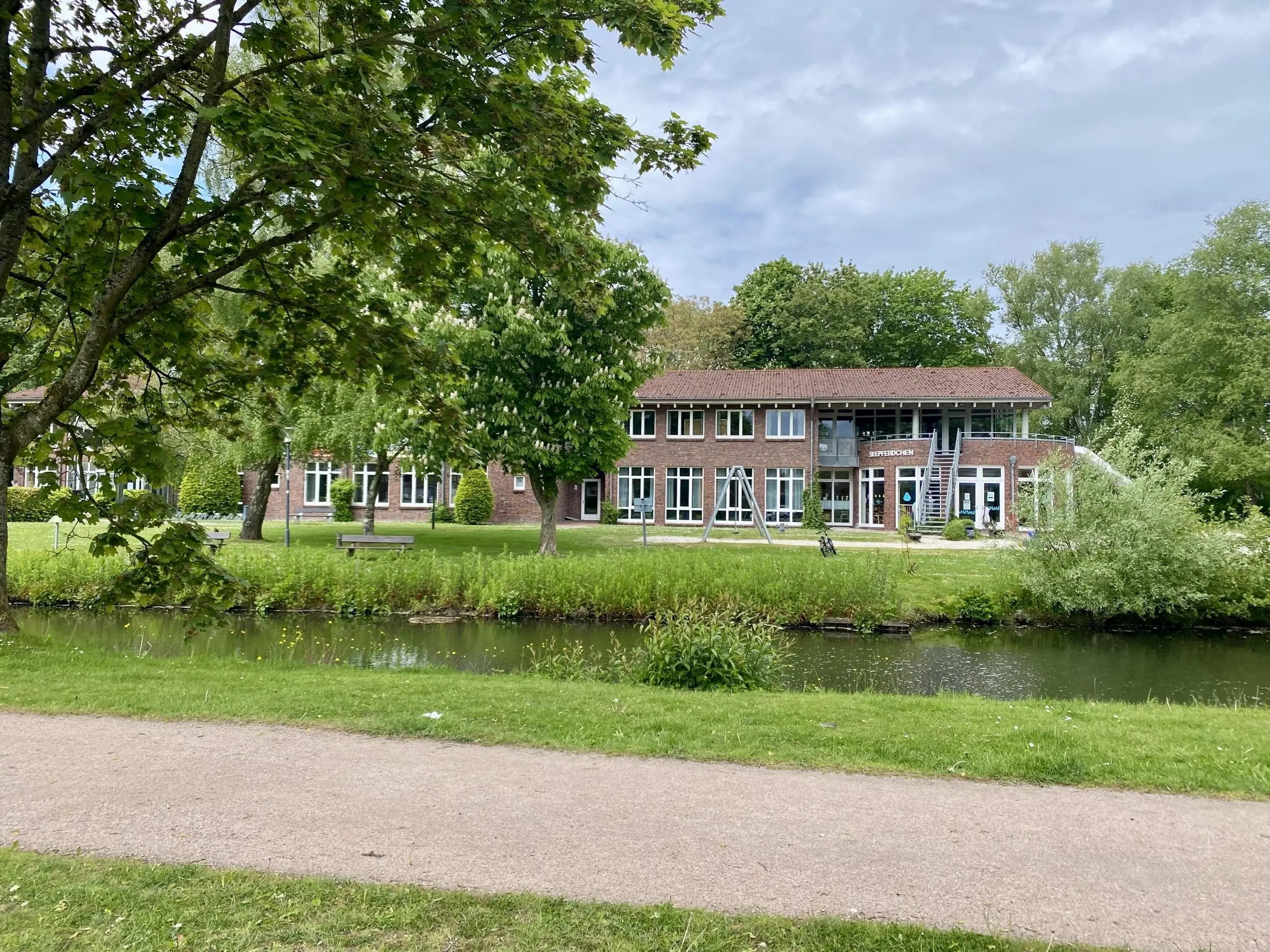 Brick school/municipal building with red-tiled roof amid a green park and trees, a small pond in the foreground.