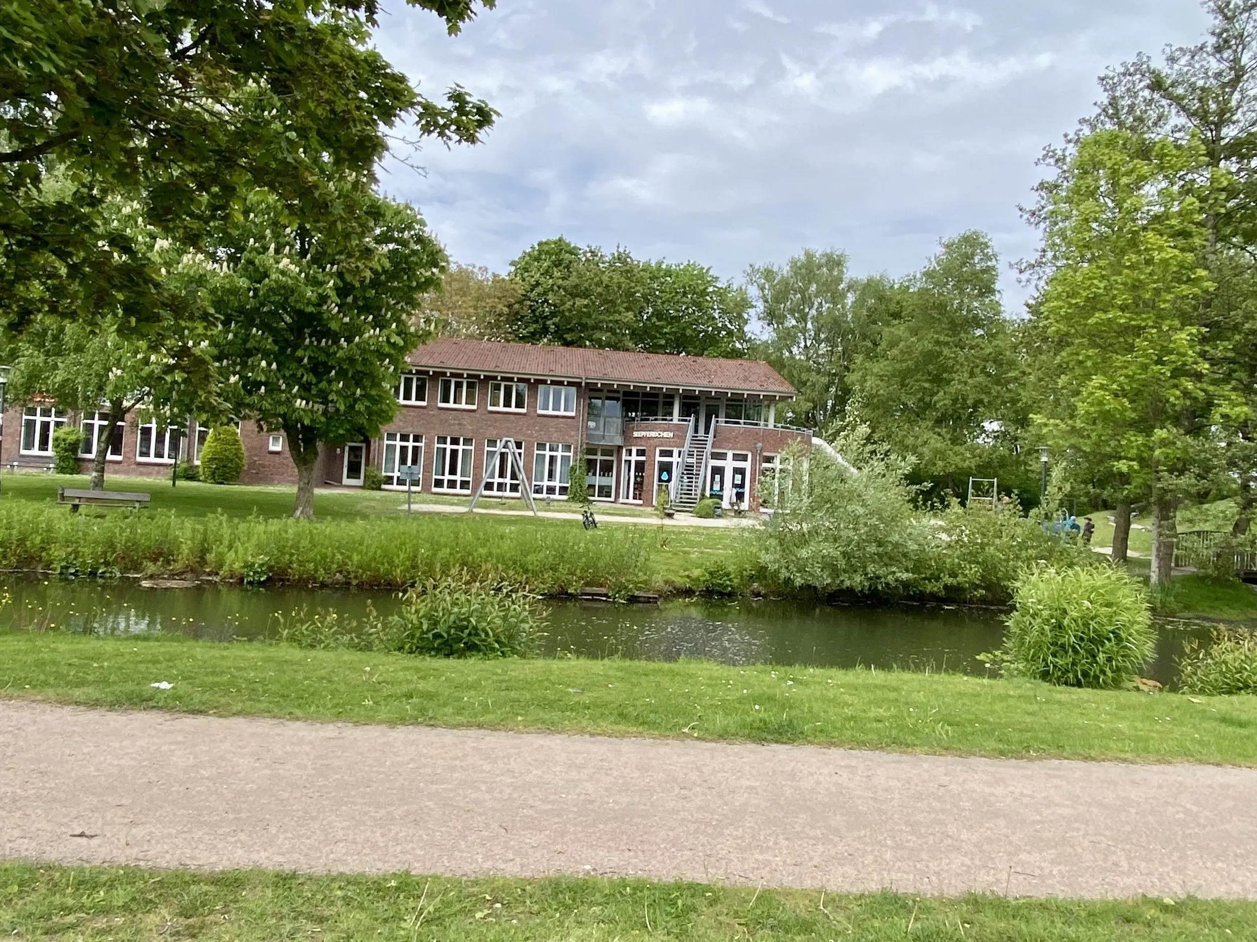 Brick building with large windows beside a pond, framed by green trees in a sunny park.