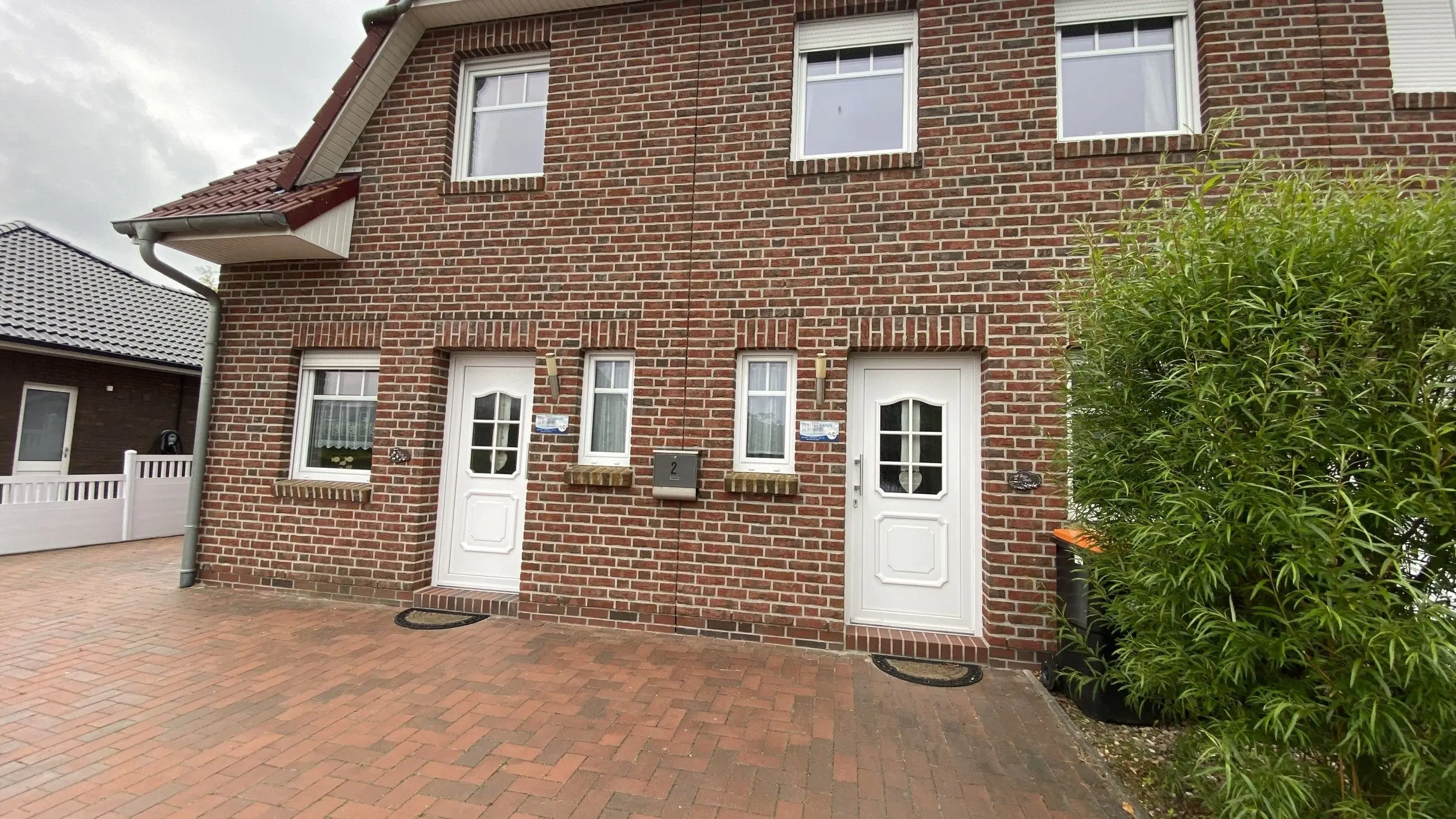 Brick townhouse façade with two white doors and ground-floor windows, a paved brick walkway, and a tall green shrub on the right side.
