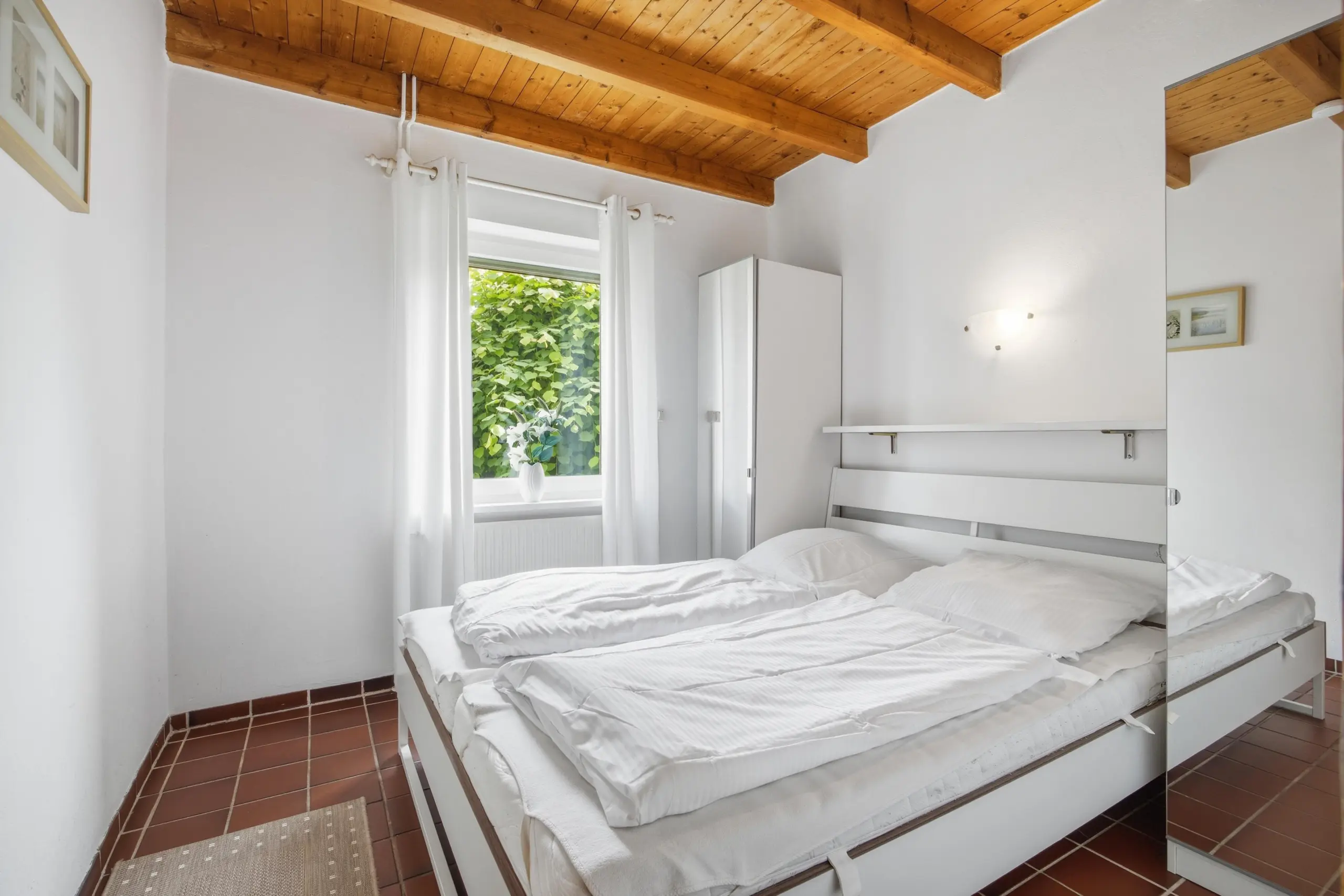 Bright white bedroom with exposed wooden ceiling beams, two beds pushed together, and white curtains by a window showing green foliage outside.