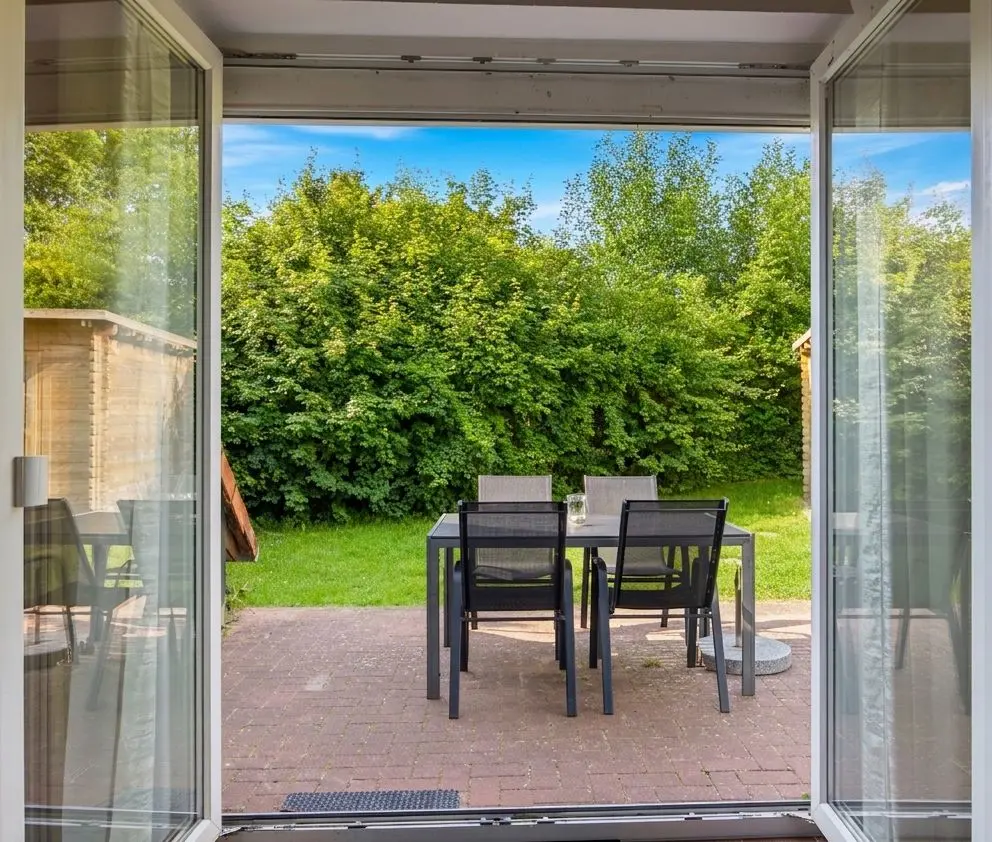 Outdoor dining table with six chairs on a brick patio, viewed through open glass doors to a hedge and lawn.
