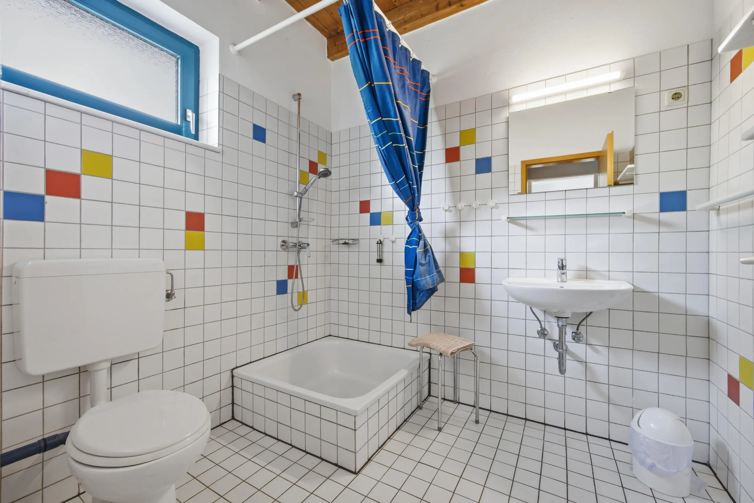 Bright white-tiled bathroom with a blue-framed window, colorful tile accents, and a blue striped shower curtain near a bathtub. That same tile pattern continues on the walls and floor, with a wall-mounted sink and mirror visible above.