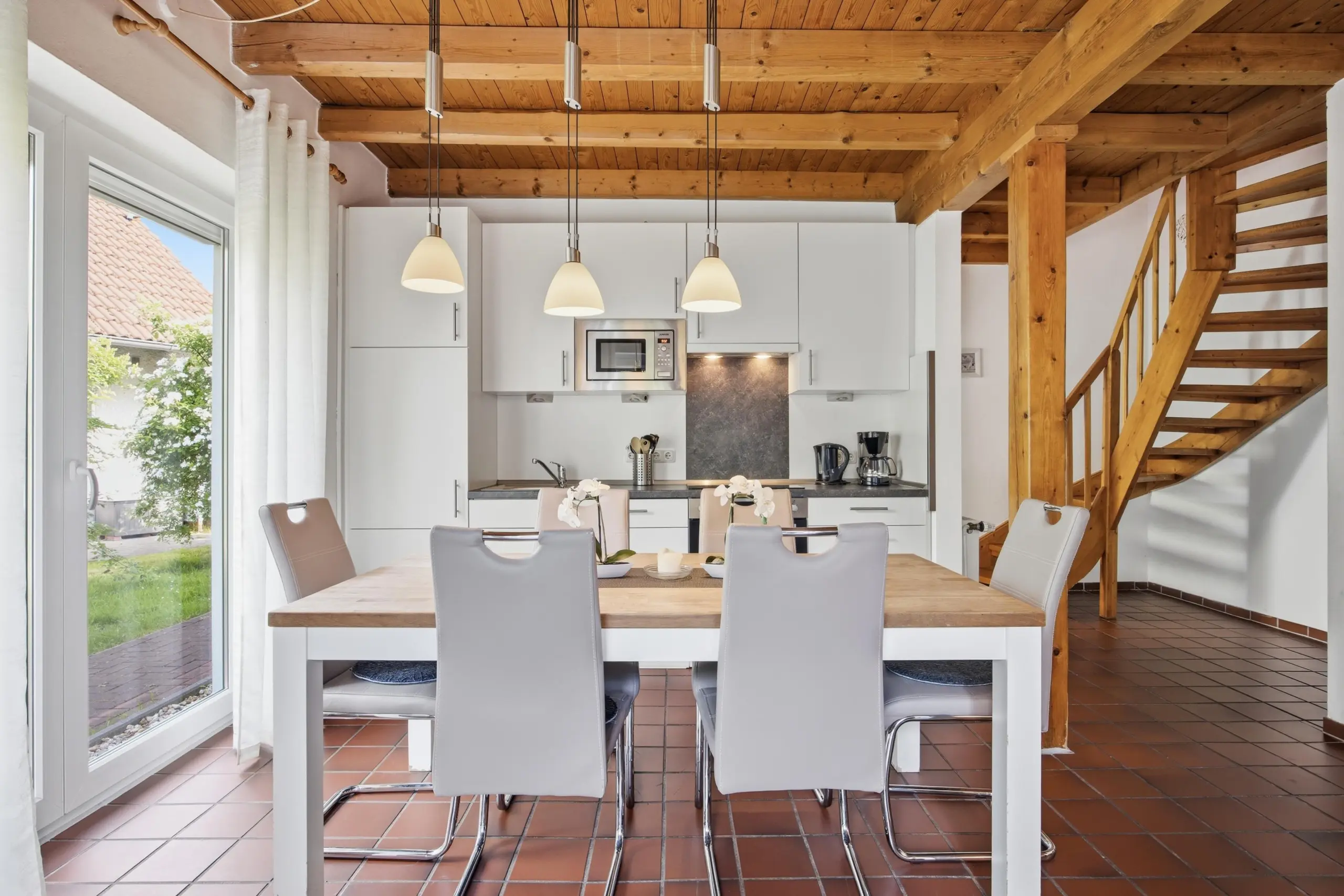 Bright kitchen-dining area with white cabinets, wooden beams, and three pendant lights above a large wooden table. Sliding glass door to garden on left.