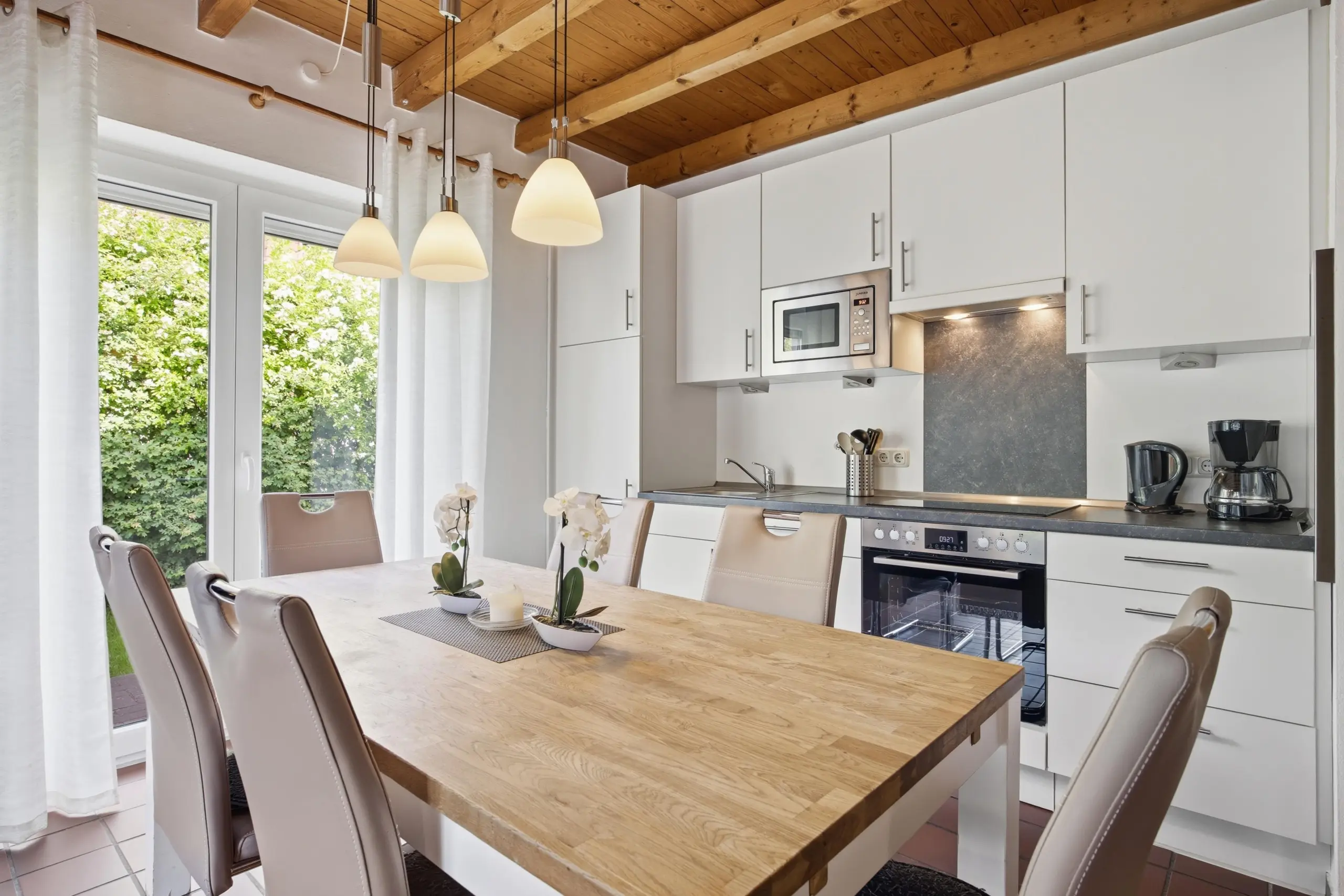 Modern white kitchen with wooden ceiling beams, pendant lights, and a wooden dining table set for four near large windows.