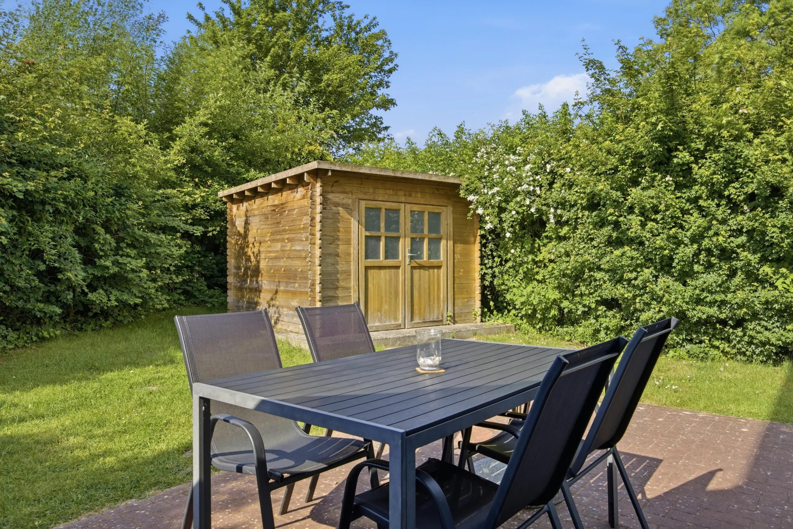 Wooden garden shed surrounded by dense green hedges and trees, with an outdoor dining table and chairs in the foreground under a blue sky.