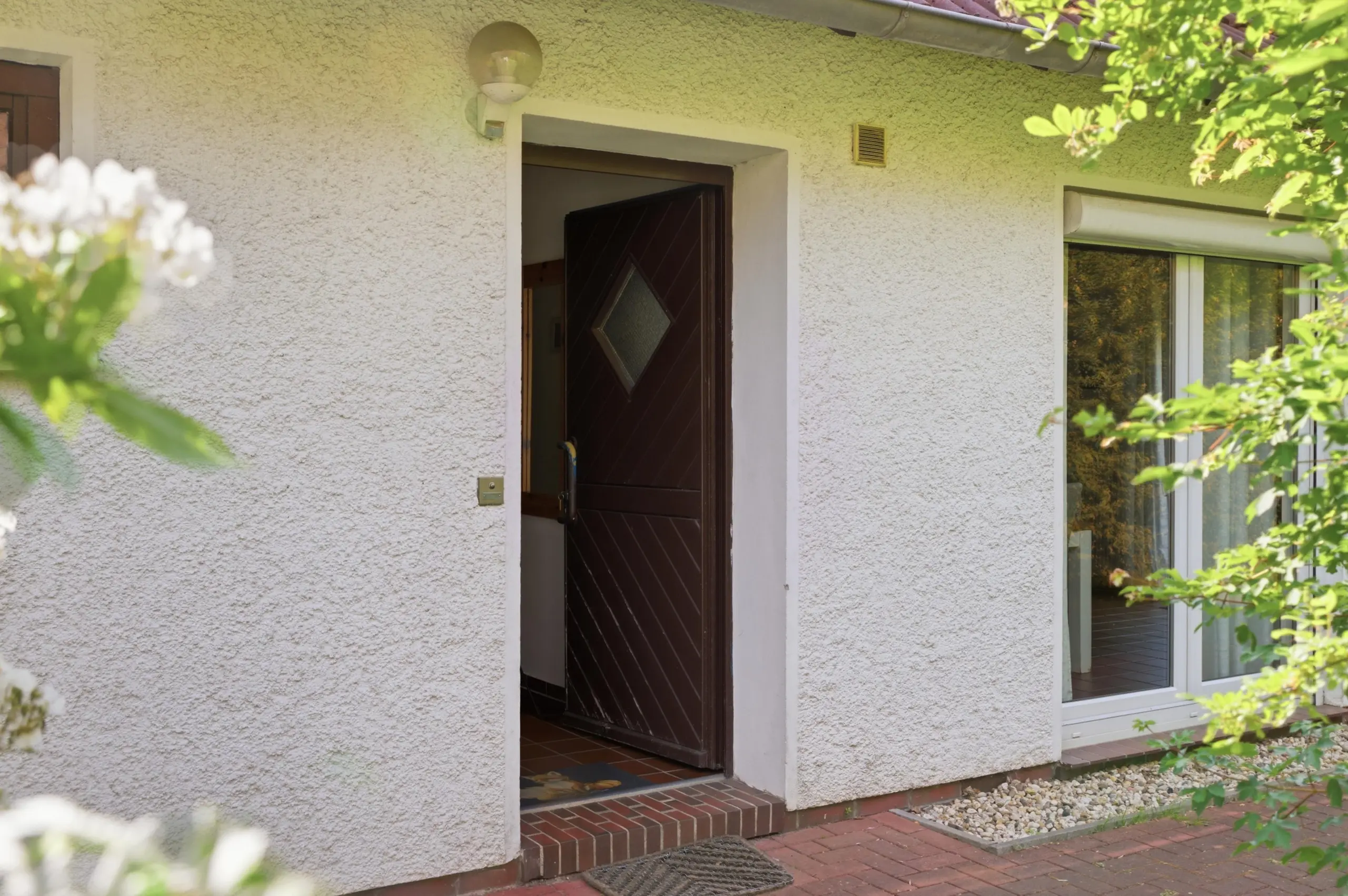 Front entrance of a beige stucco house with a dark brown door featuring a diamond-shaped window, brick step, and a nearby window with blinds.