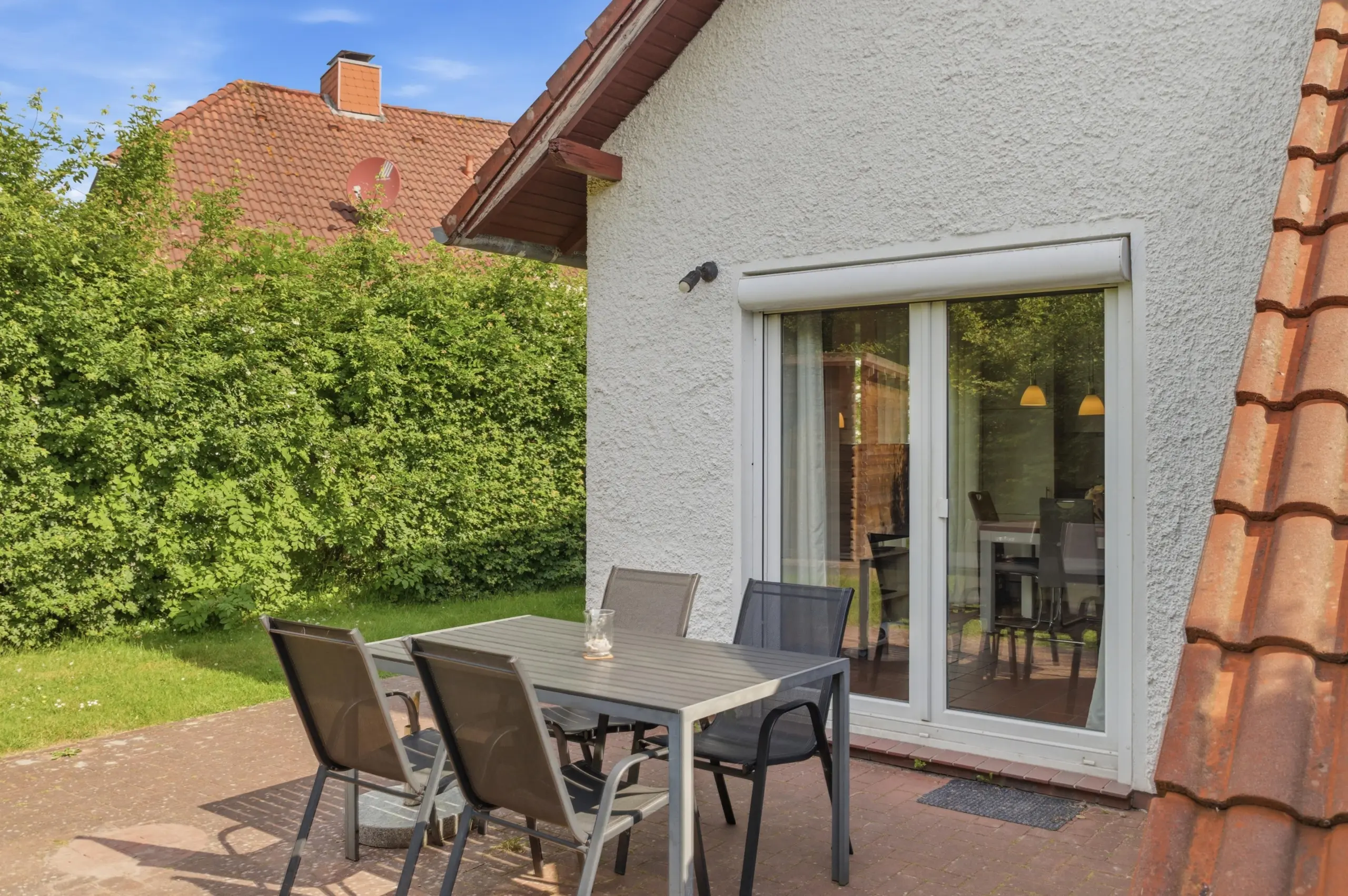 Outdoor patio with a metal dining table and six chairs beside a sliding glass door, bordered by a green hedge and a red-tiled roof.