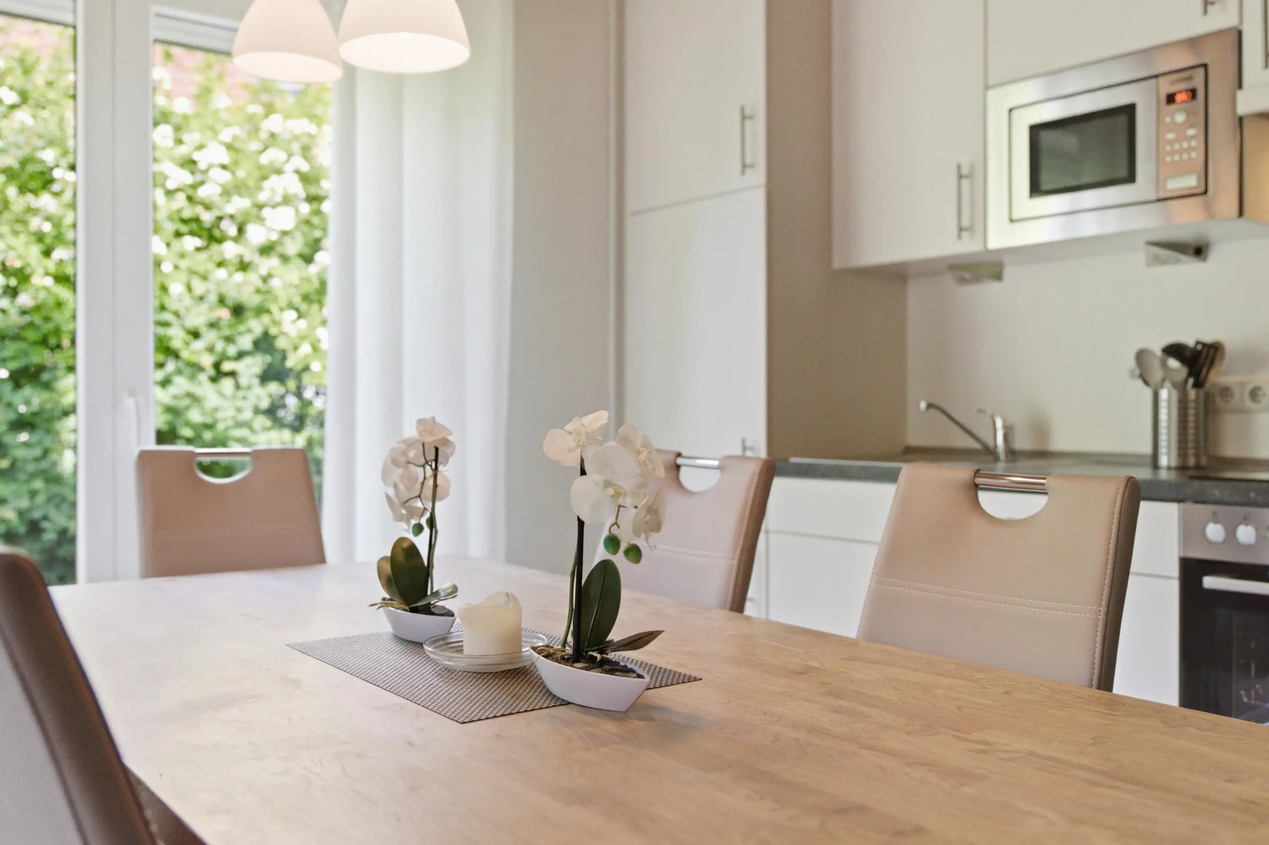 Bright kitchen-dining area with a marble table, beige upholstered chairs, and white orchid centerpieces; a stainless microwave is mounted on the wall.