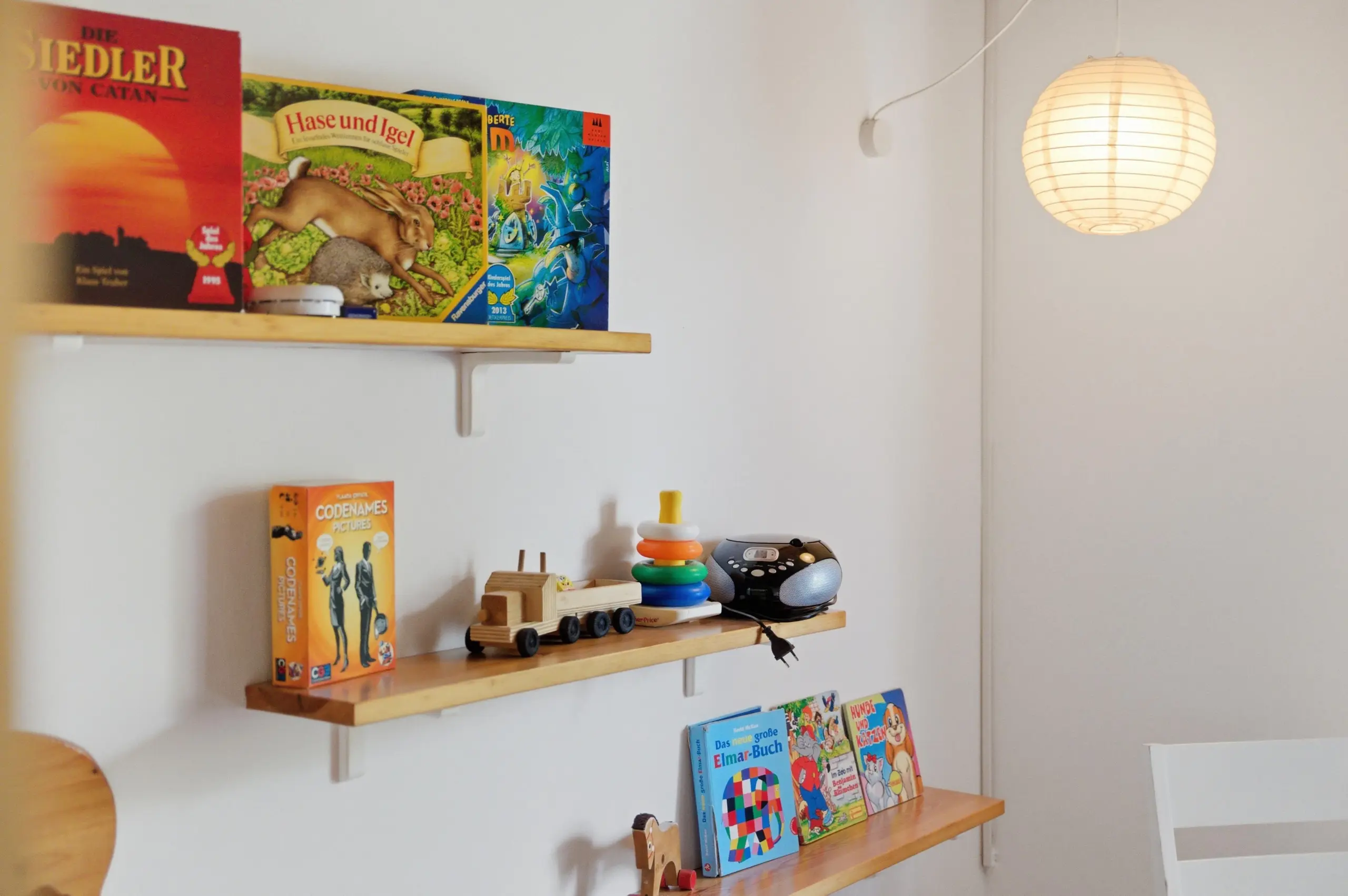 Wall-mounted shelves hold colorful children's books and wooden toys, with a glowing paper lantern lamp to the right.