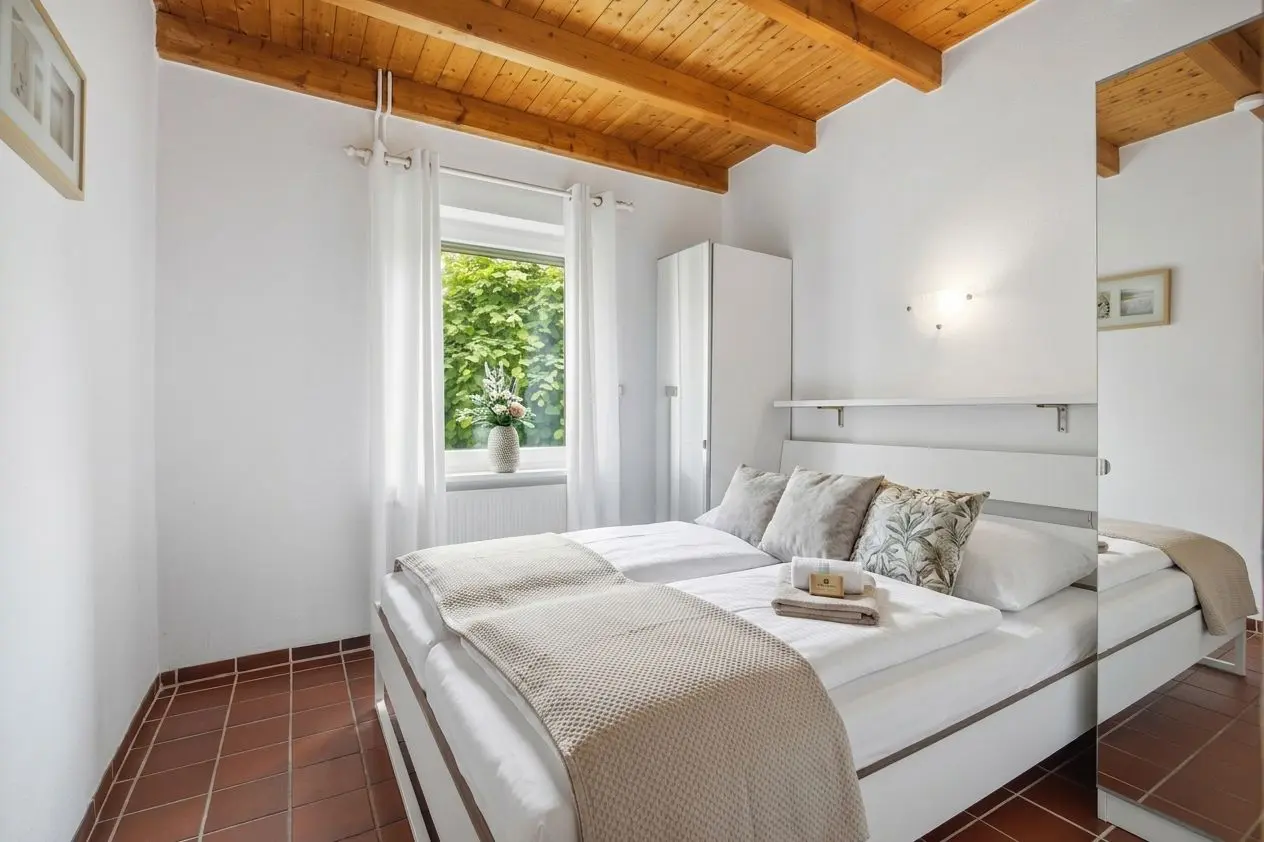 Bright bedroom with white walls, wooden beam ceiling, and terracotta tiled floor; double bed with beige throw and decorative cushions opposite a mirrored wardrobe.