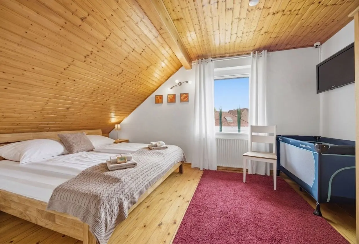 Attic bedroom with a wooden sloped ceiling, a large bed, neutral throws, and a red rug underfoot next to a window with white curtains.