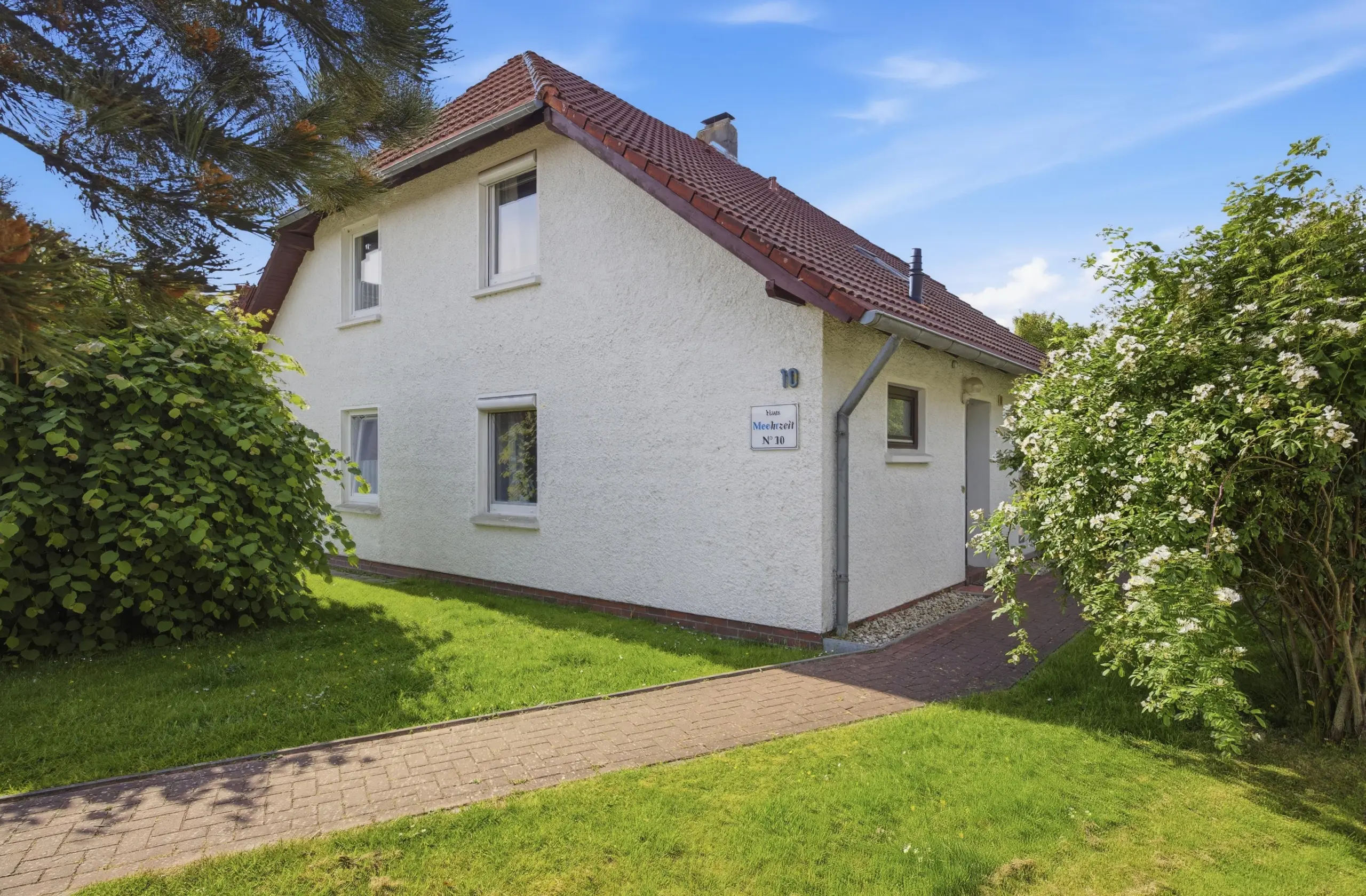 White stucco house with a red-tiled roof, flanked by green bushes and a brick walkway on a sunny day.