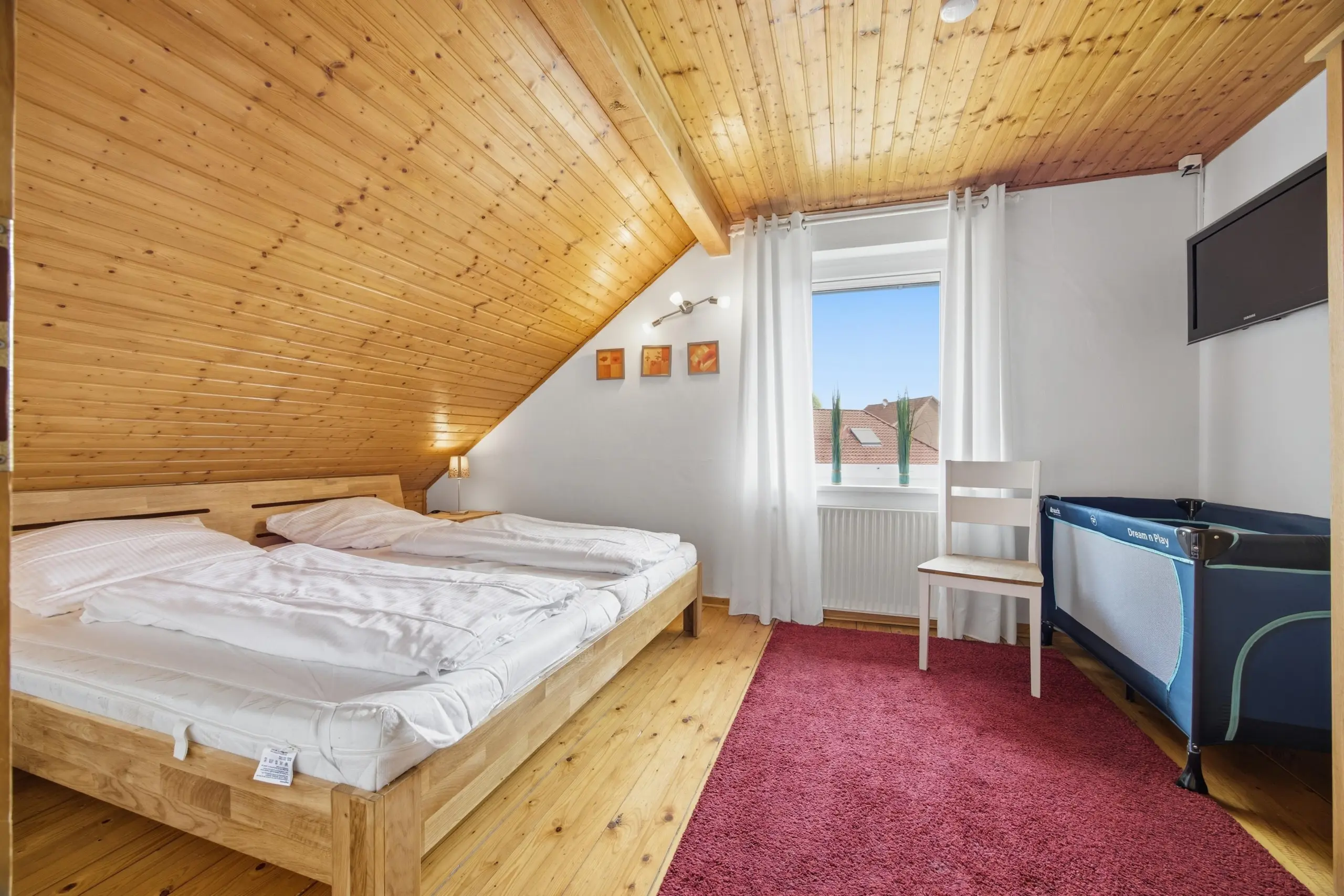 Attic bedroom with a slanted wooden ceiling, two single beds, white bedding, and a red rug; window with white curtains and wall art.