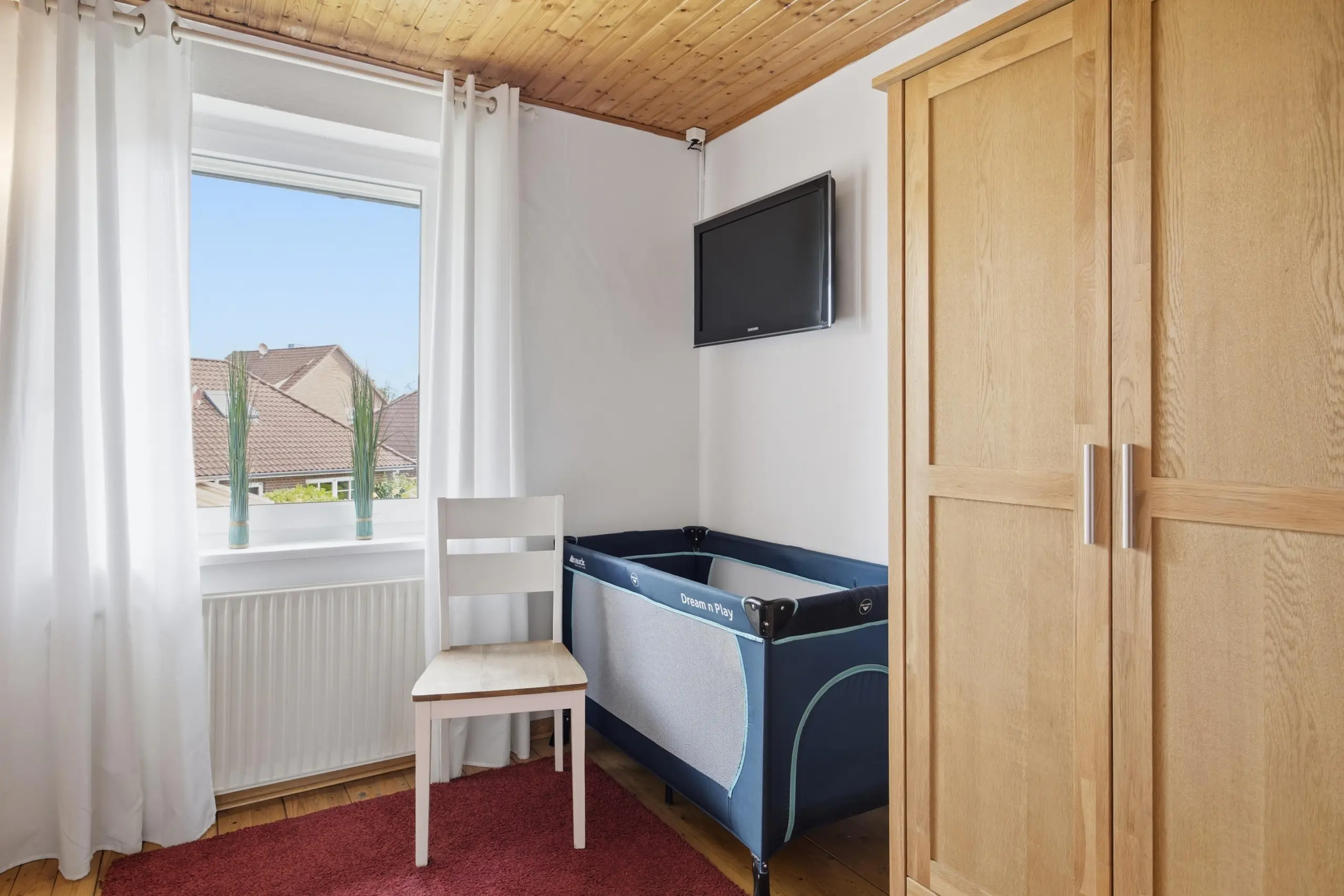 Bright corner of a bedroom with a wall-mounted TV, a blue playpen, white curtains by a window, and a tall wooden wardrobe.
