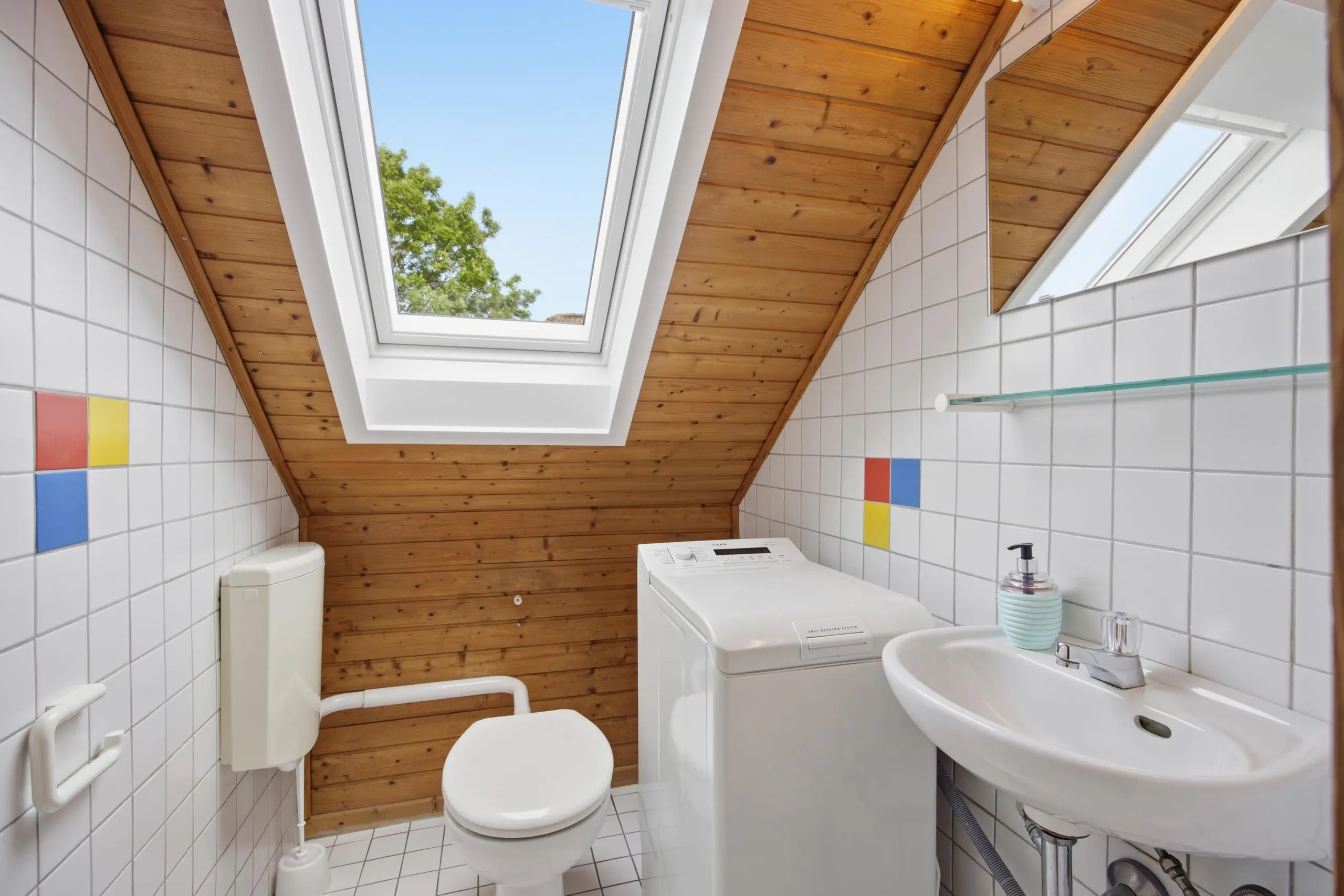 Bright bathroom with white tiled walls, a sloped wooden ceiling, and a skylight showing blue sky and a tree; toilet, sink, and washing machine visible.