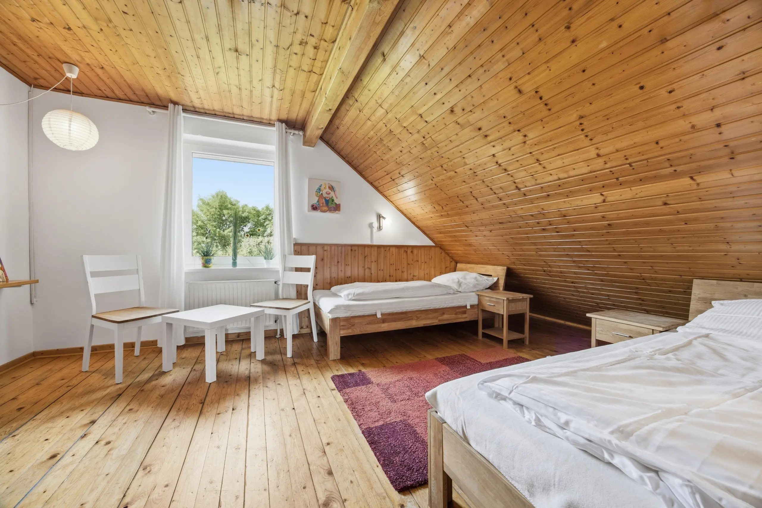 Attic bedroom with pine slatted ceiling, two single beds, white furniture, and a window letting in sunlight.