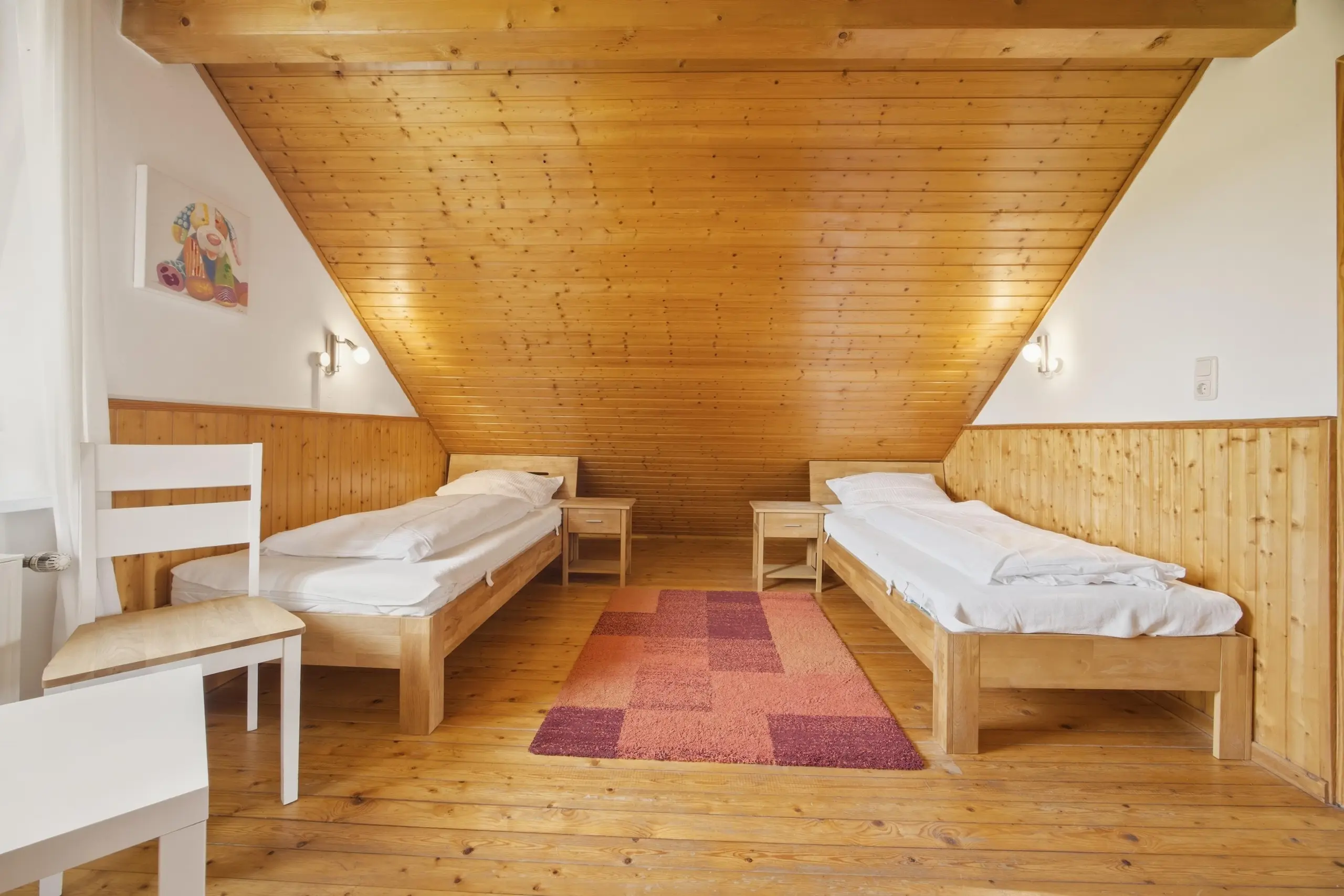 Cozy attic bedroom with two single wooden beds facing each other, white bedding, and a pink area rug between them under a slanted wooden ceiling.
