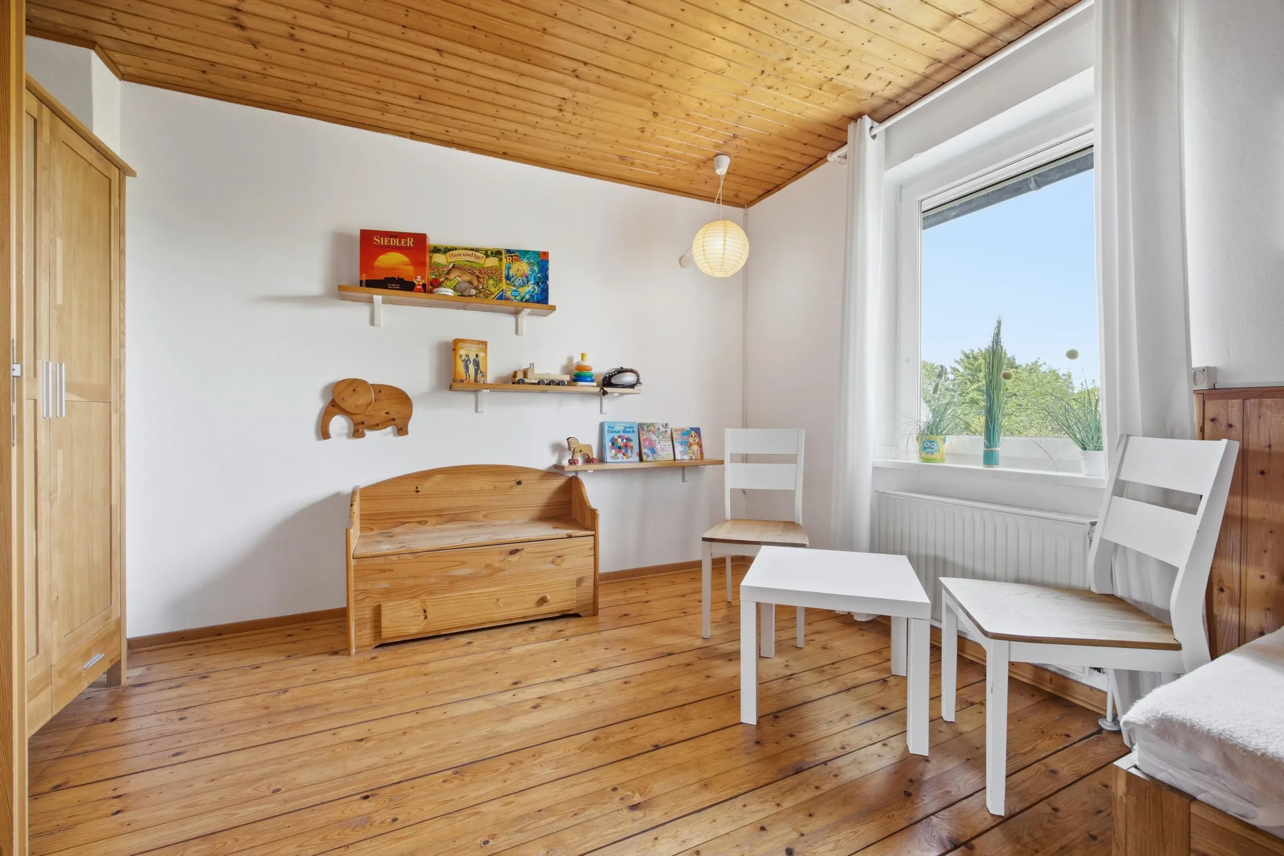 Bright children's playroom with wooden floor, ceiling, and white walls, featuring wall shelves of books and toys and a wooden bench beneath them.